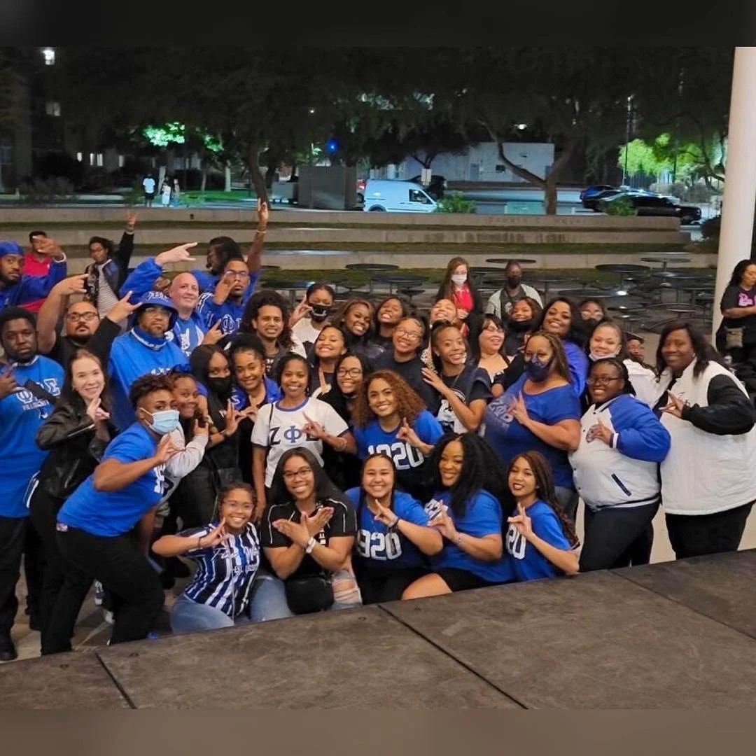 Group of smiling women and men, some wearing Sigma Phi Beta apparel, gathered outdoors at night, posing for a group photo.