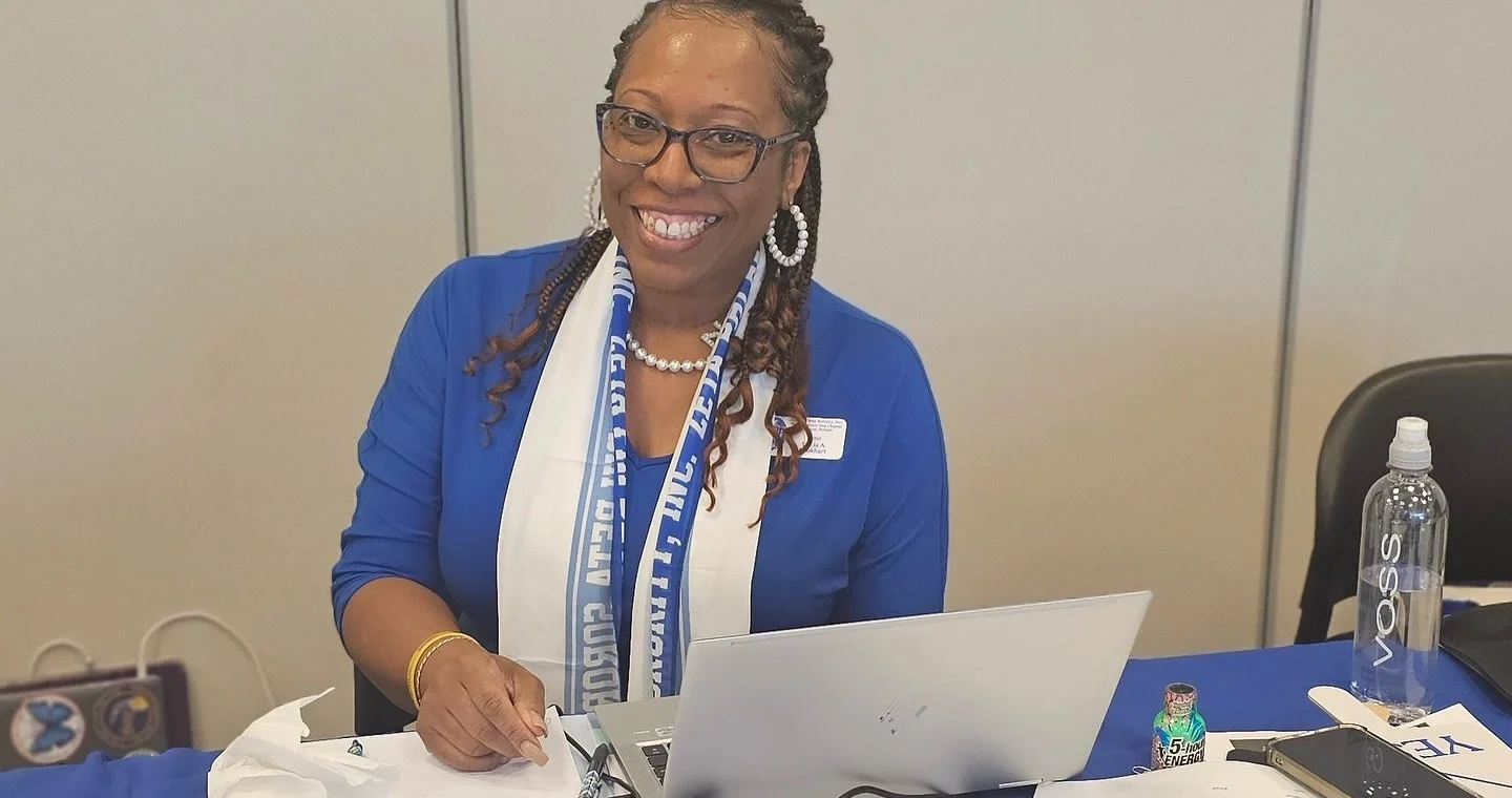 Smiling woman with glasses, wearing a blue jacket and a white and blue scarf, sitting at a table with a laptop, water bottle, and other items, in an indoor setting.