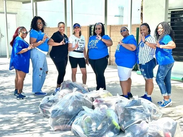 A group of eight women standing outside on a sunny day, posing beside several large trash bags filled with collected items, suggesting a community cleanup event.