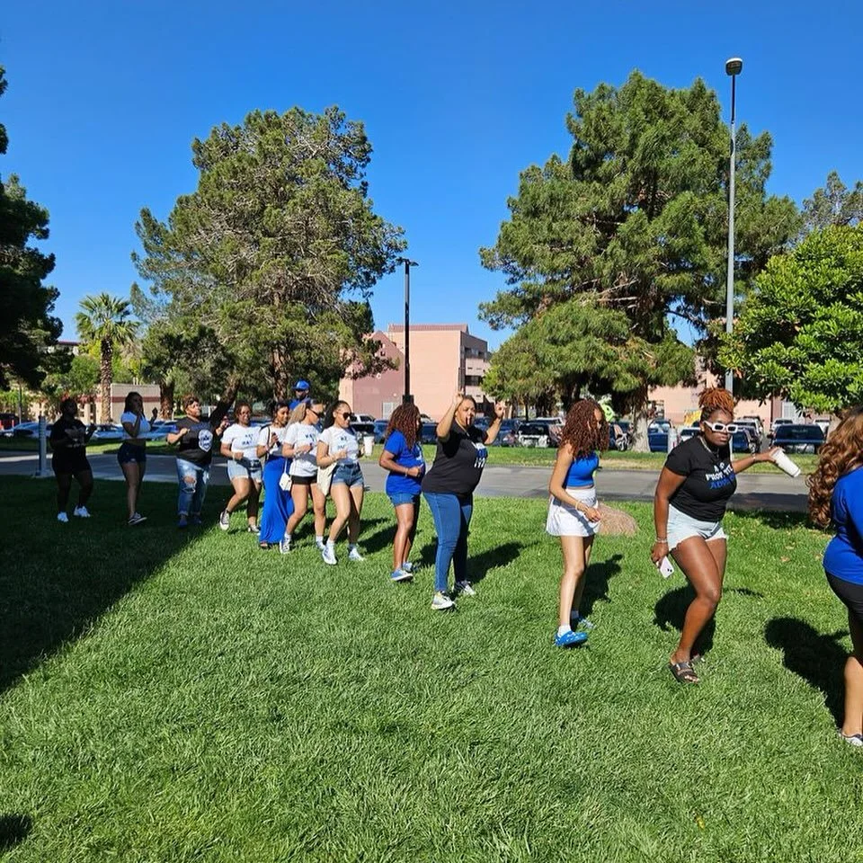 A group of women standing and sitting in a grassy park area, participating in an outdoor event on a sunny day. Some women are wearing blue and white clothing, and they are organized in a line or circle, with trees and buildings in the background.