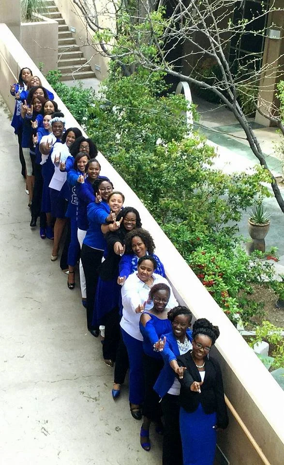 A line of women standing along a balcony railing, pointing towards the camera, with a garden and trees in the background.