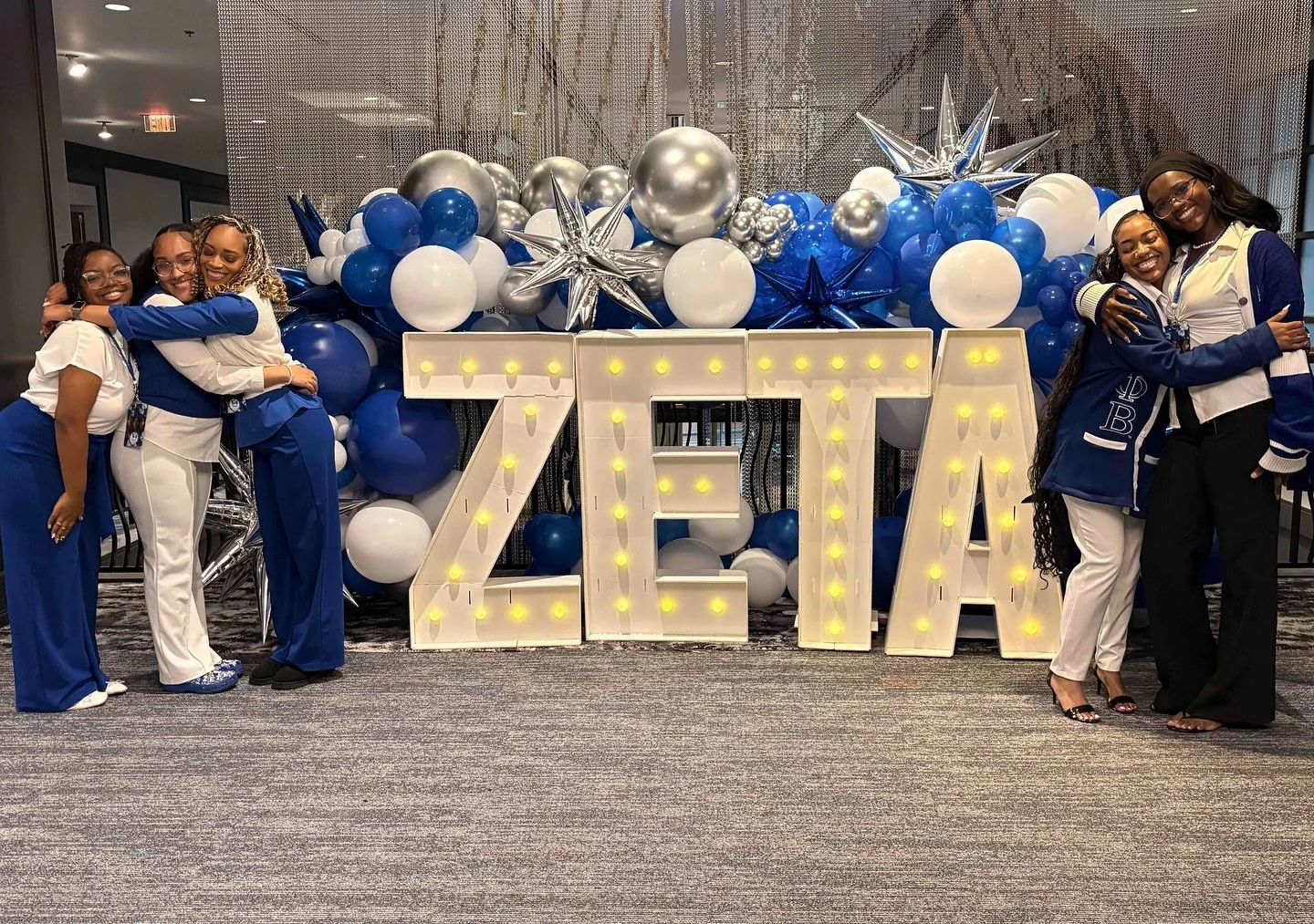 Six women are hugging and smiling in front of a large illuminated sign that reads 'ZETA'. Behind them, there is a decorative backdrop of blue, white, and silver balloons and star-shaped balloons.