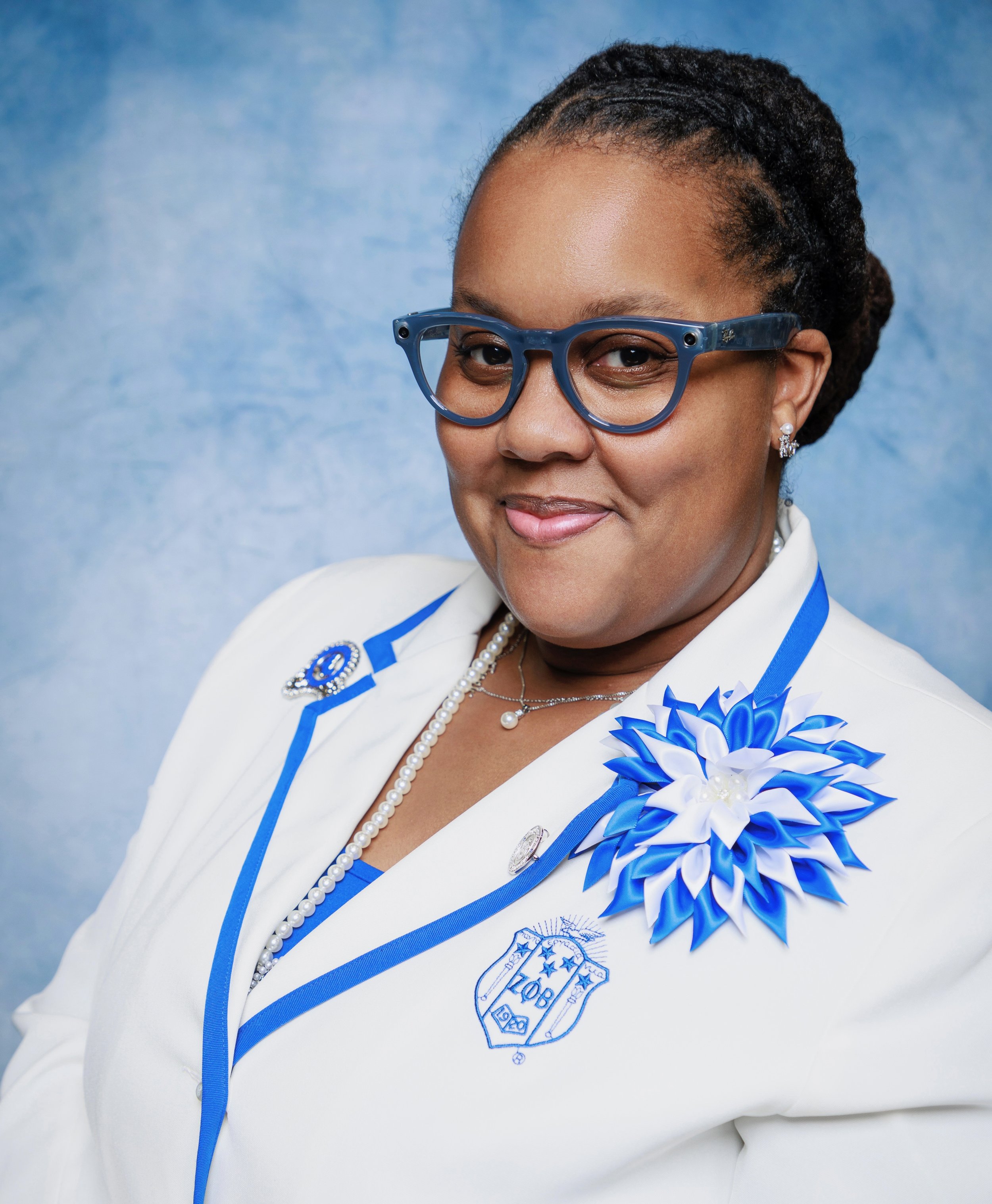 A woman wearing glasses, a white blazer with blue accents, and a large blue and white ribbon pin on her blazer. She has braided hair and is smiling with a blue textured background.