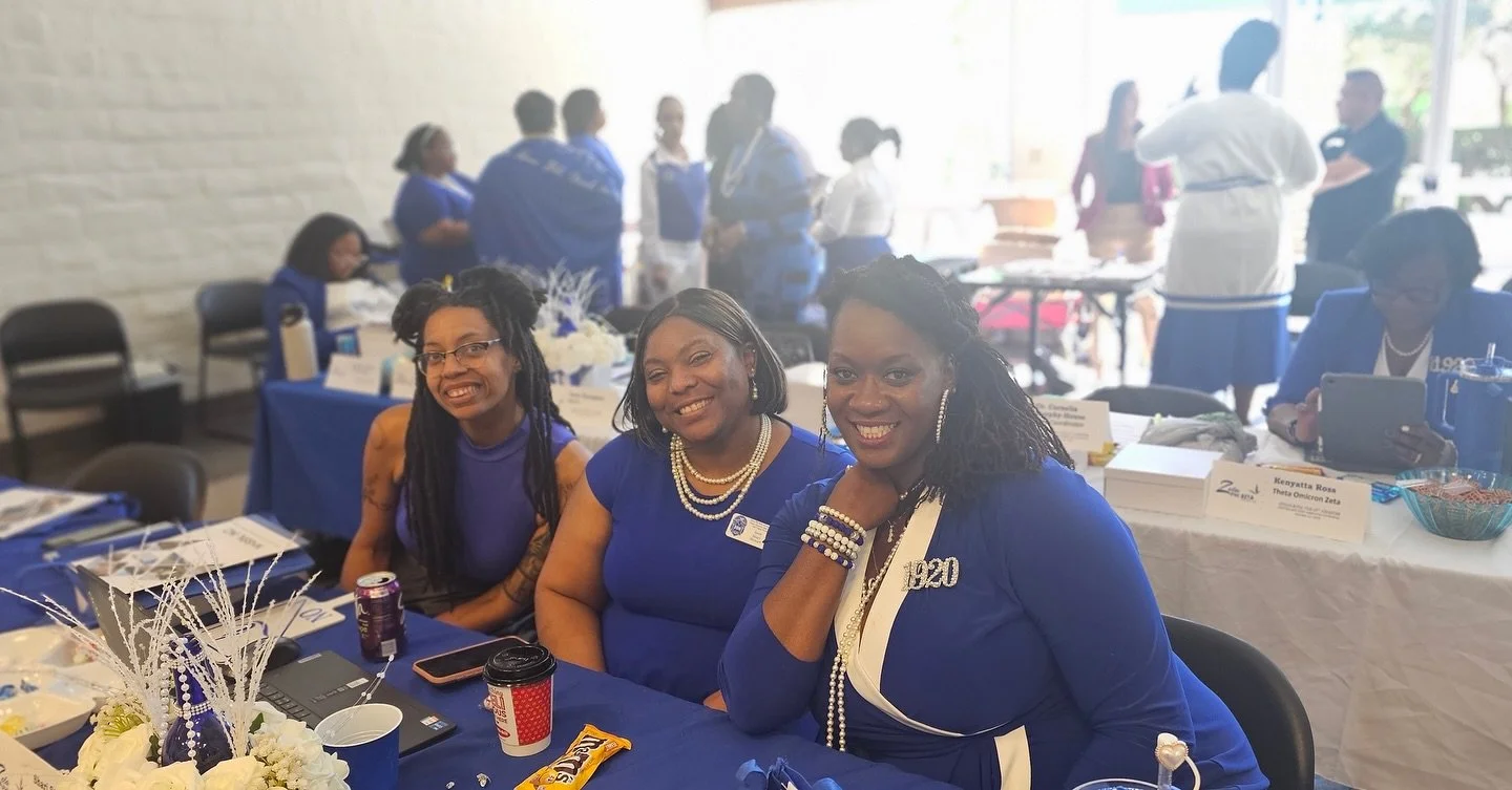 Three women wearing blue dresses sitting at a table during a social event or conference, smiling at the camera, with other people in the background.