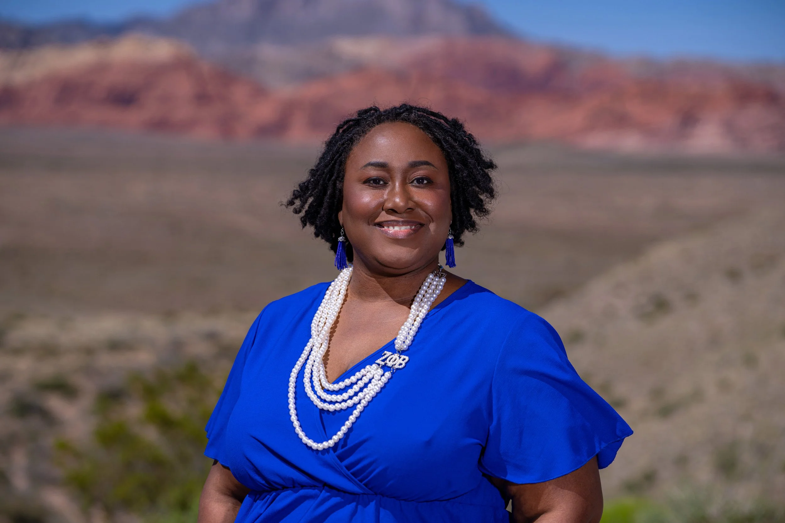 A woman in a bright blue dress and jewelry standing outdoors with a desert landscape and red rock formations in the background.