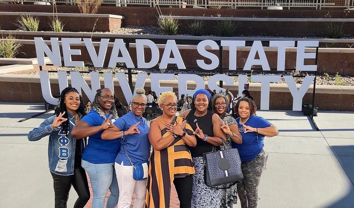 Group of diverse women standing in front of a Nevada State University sign, posing and smiling for the camera.