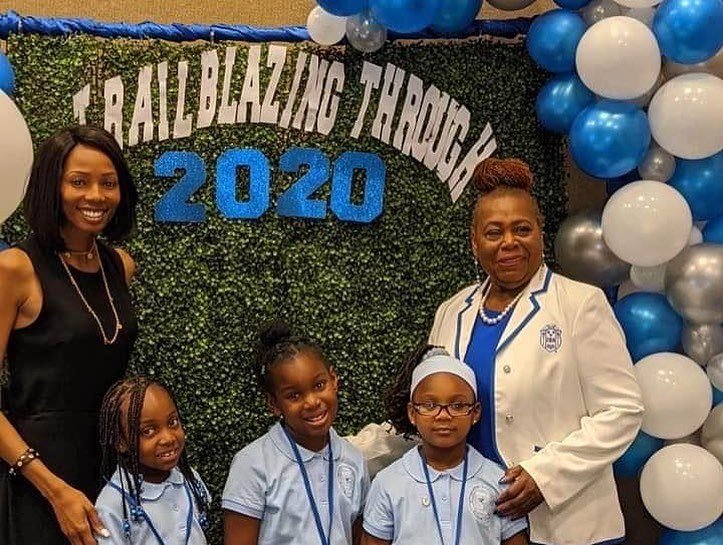 Group photo of two adult women and three young girls at a school event with a backdrop reading 'Trailblazing Throne 2020' and decorated with blue, white, gray, and black balloons.