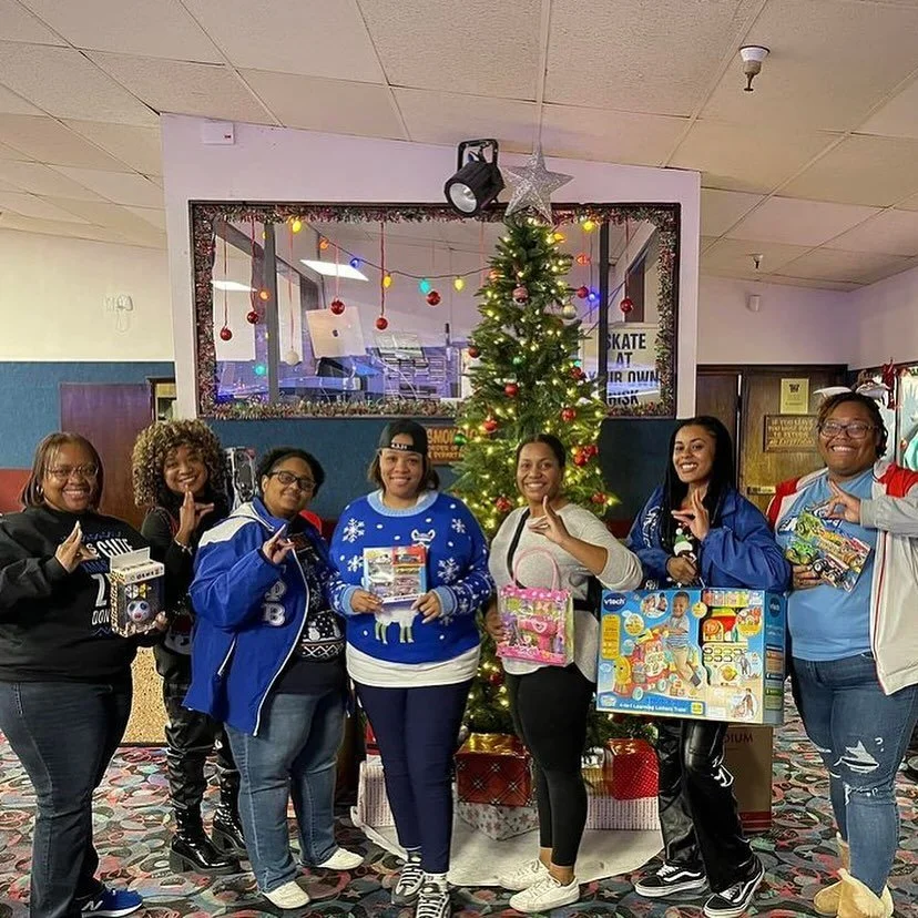 Group of women posing in front of a decorated Christmas tree, holding toys and gifts, in a festive indoor setting.