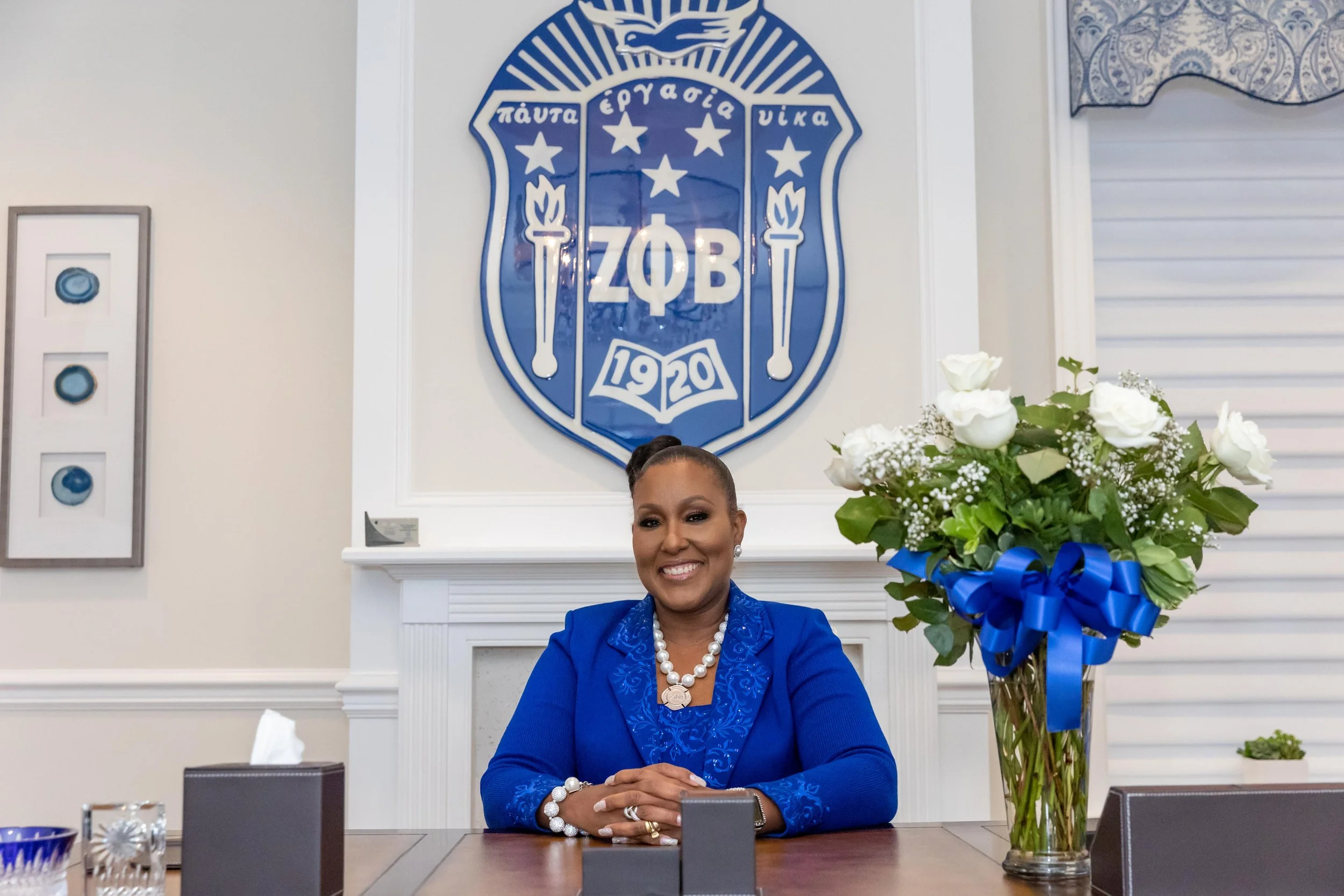 A woman sitting at a table smiling, in front of a large blue and white Greek organization emblem on the wall, with a tall floral arrangement of white roses and greenery with a blue ribbon.