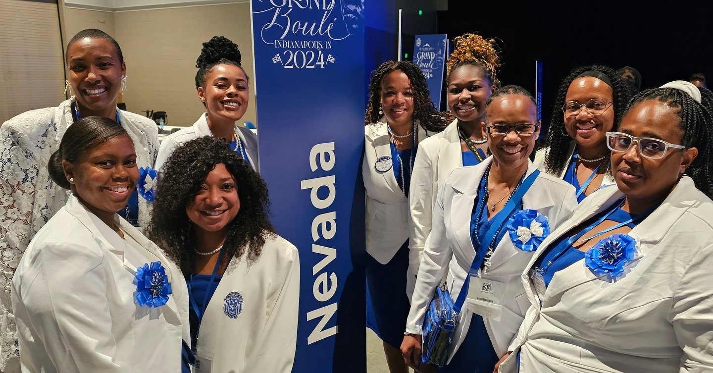 Group of women in white coats at a professional conference, wearing blue ribbons and badges, standing beside a blue sign that says 'Nevada' and 'Grand Bowl Indianapolis 2024' in the background.