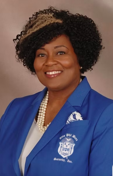 A woman with short curly hair wearing a blue blazer with a pearl necklace and a white top, smiling in a professional portrait.