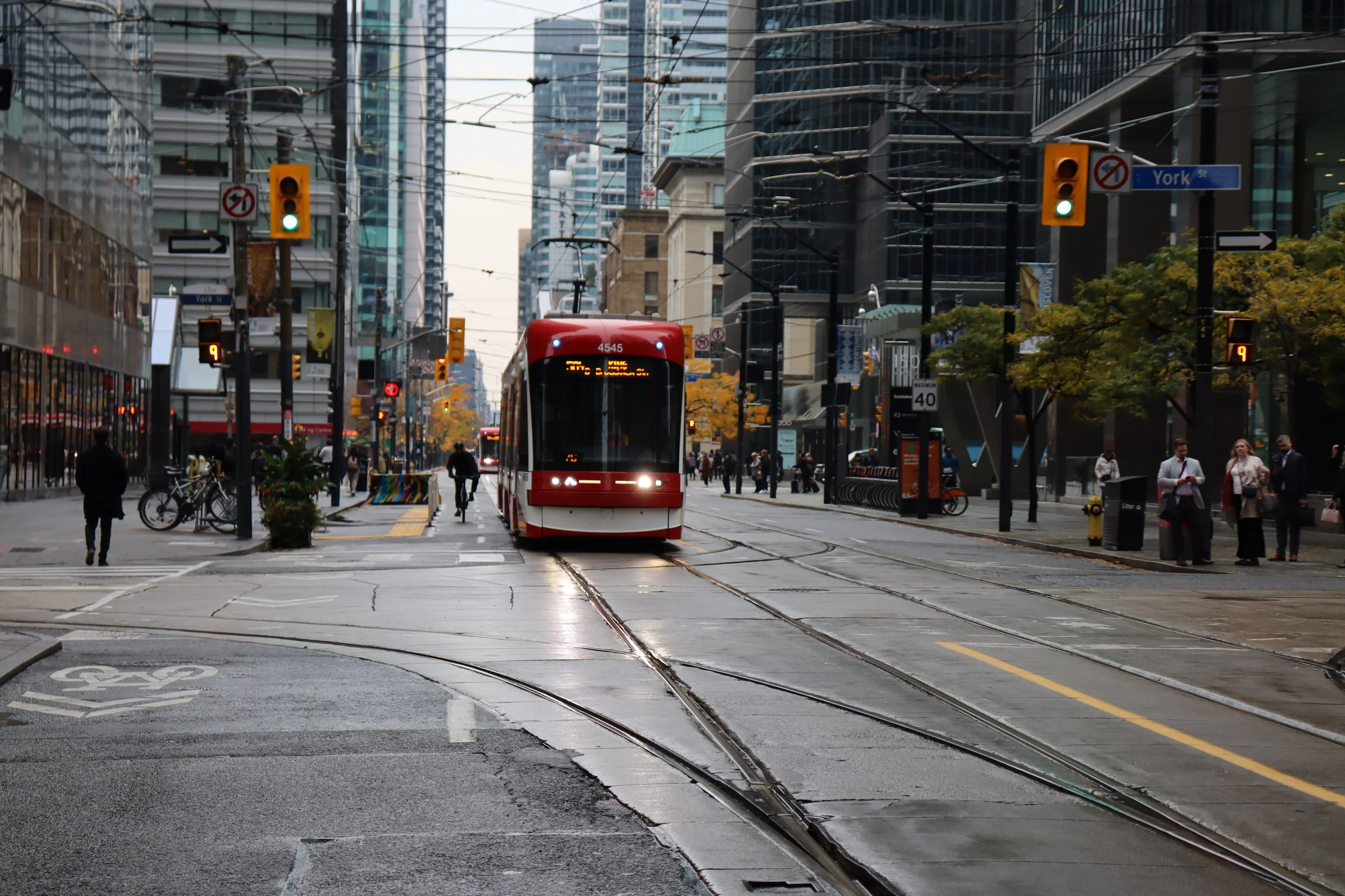 A city street scene featuring a red streetcar on tracks, with pedestrians walking on the sidewalk and buildings in the background. The street is wet, likely from rain, and there are traffic signals and signs visible.