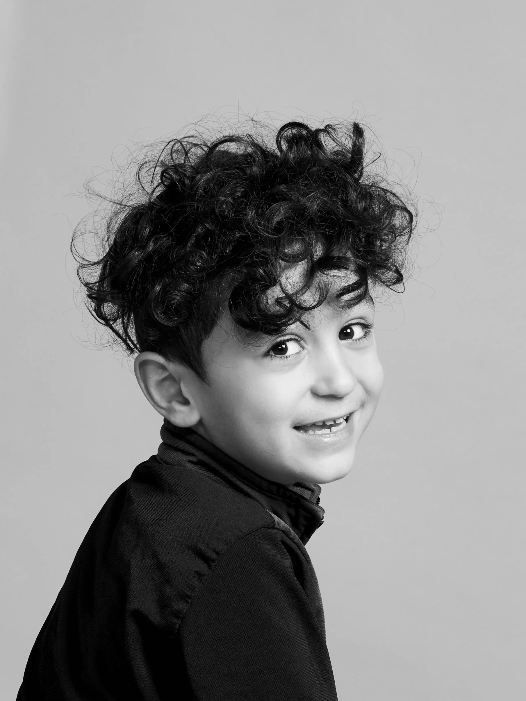 Sort-hvid portrætfotografi af et smilende barn med krøllet hår iført en sort jakke, vendt let til højre, mod kameraet.