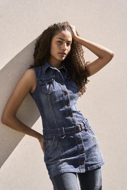 Kvinde i denimkjole og jeans står mod en lys væg, hun har krøllet hår og en hånd i håret.
