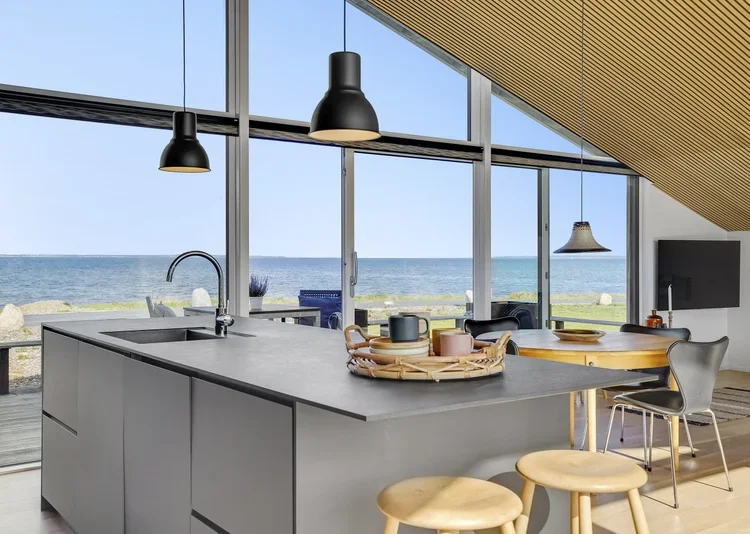 Moderne åben køkken med udsigt til havet, store vinduer, lyse trælofter, sort gasplade og spisebord med stole.