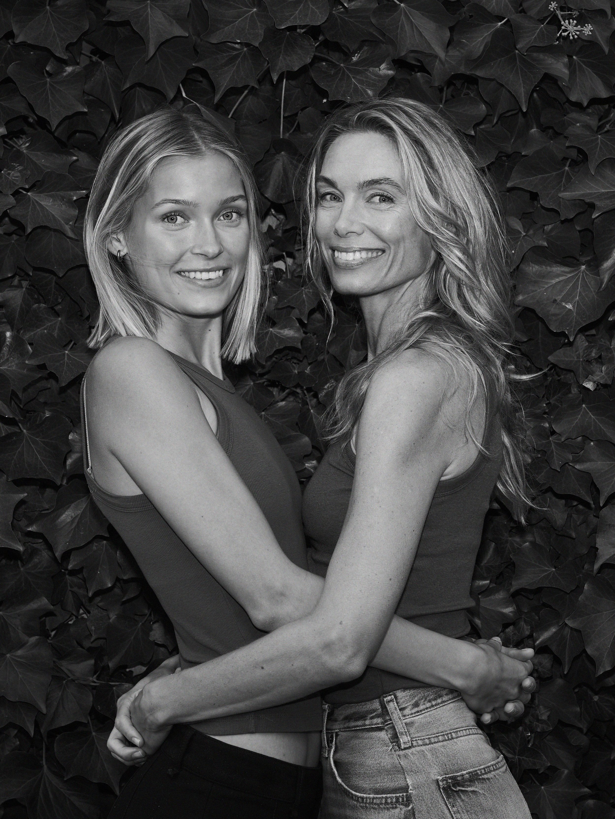 To kvinder står tæt sammen foran en væg dækket af blade, smilende mod kameraet.