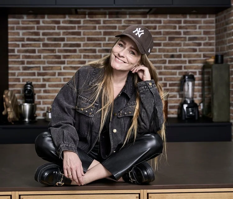 Kvinden sidder krydsbenet på et bord i et køkken, smilende med kinden mod hånden, iført en sort baseballkasket, sort denimjakke og læderbukser.