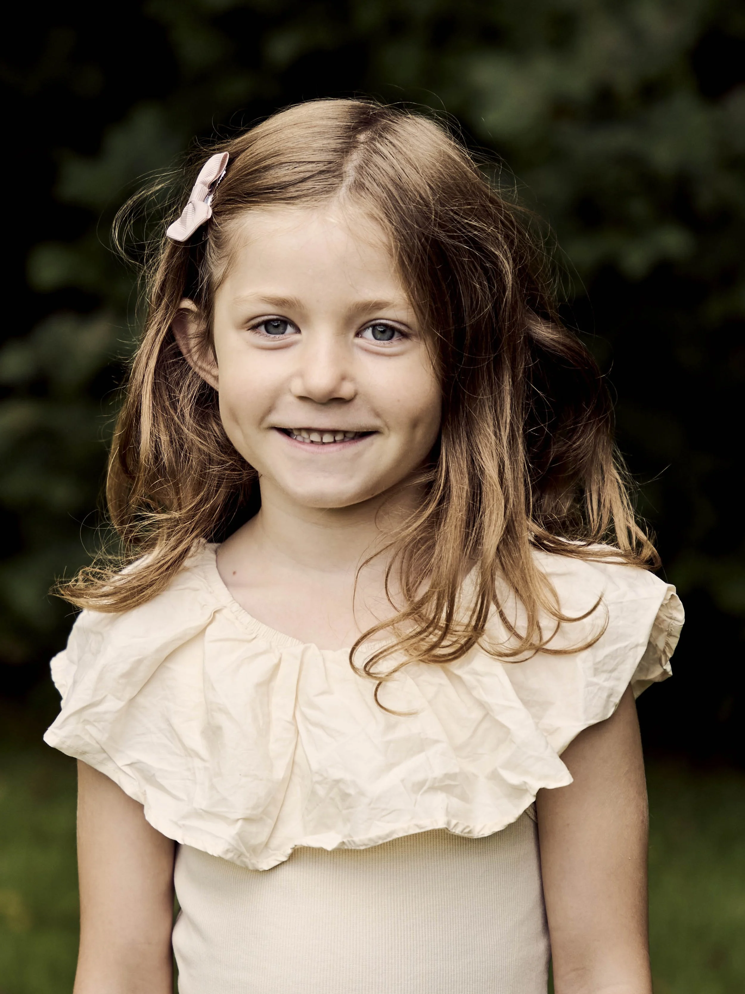 Smilende ung pige med brunt hår og blå øjne, iført beige kjole med store blomsterfalder rundt halsen, udendørs med grønne træer i baggrunden.