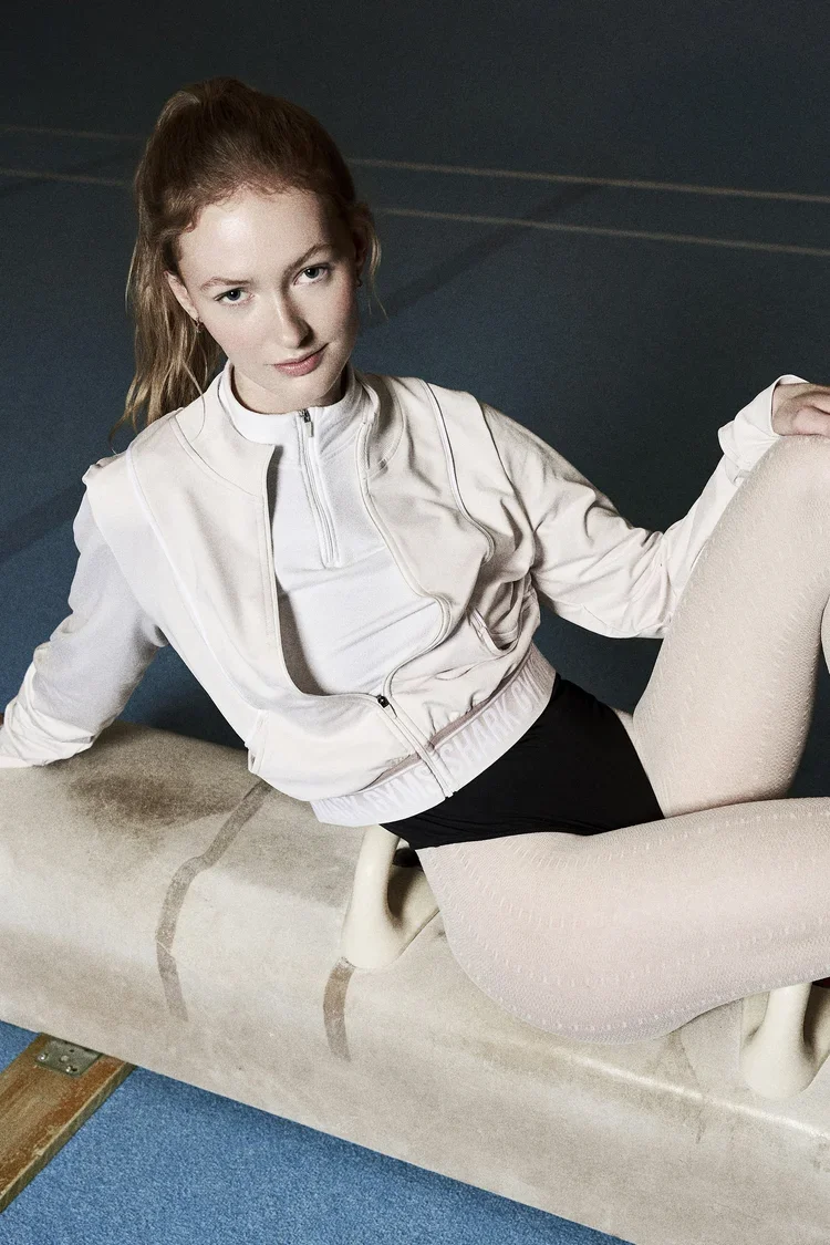 Kvindelig fitnessudøver sidder på en ergonomisk træningsmaskine i træningscenter iført lyserødt træningstøj og hvide leggings. Model. Esther Knudsen. Modelbureau LESS Models. Fotograf Pia Bloch