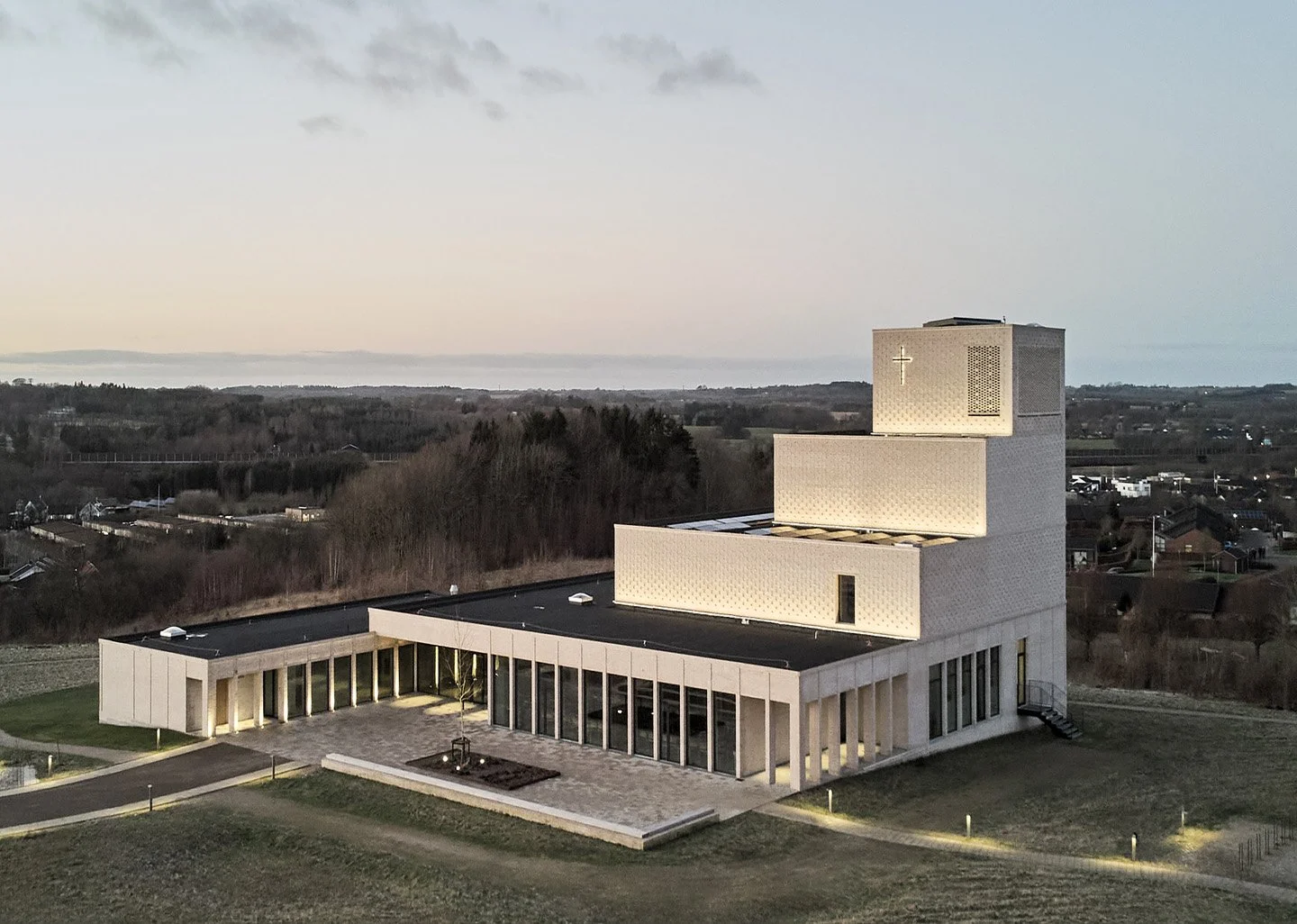 Moderne kirke med lyse facader og kors foran, omgivet af grønne arealer ved skov og huse i baggrunden, optaget ved skumring.
