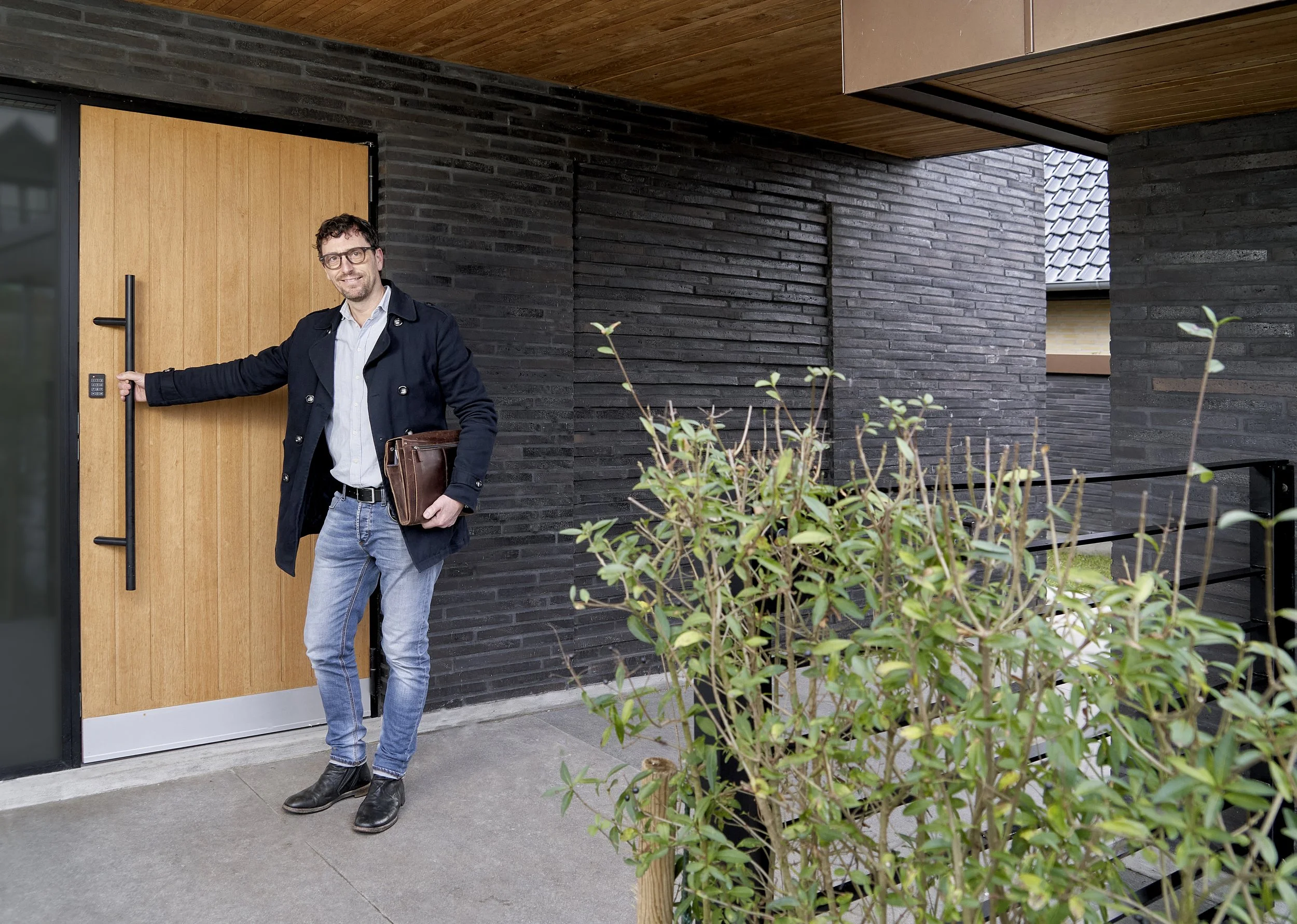 Mand står foran en sort murstensfacade og en trædør, smiler, holder en mappe, og har en jakke på.