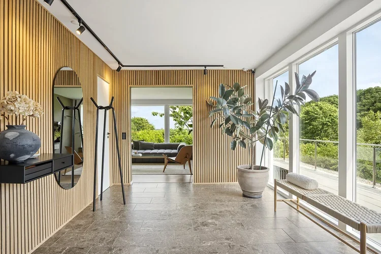 Indretning af moderne lejlighed med trægavle væg, søjler, store vinduer med udsigt til grøn natur, plante i stor potte, spejl og bænke i rattan.
