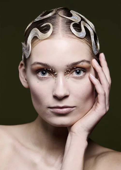 Portræt af en ung kvinde med blonde hår sat i skulpturede bølger og rhinestones omkring øjnene,, rører sit ansigt med hånden.