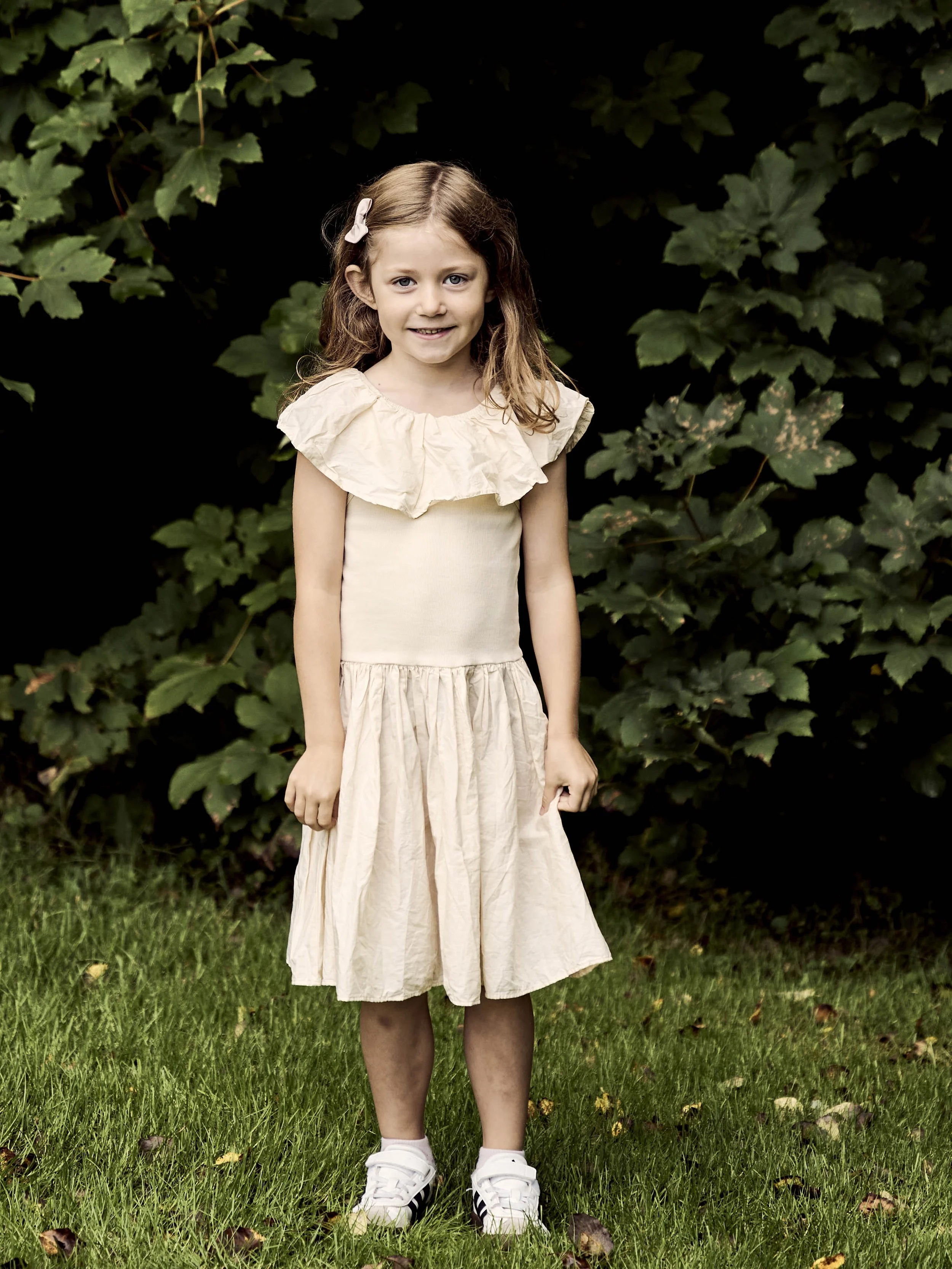 En smilende ung pige med brunt, bølget hår og en hvid skinnende kjole med store ærmer står på græs foran grønne buske.