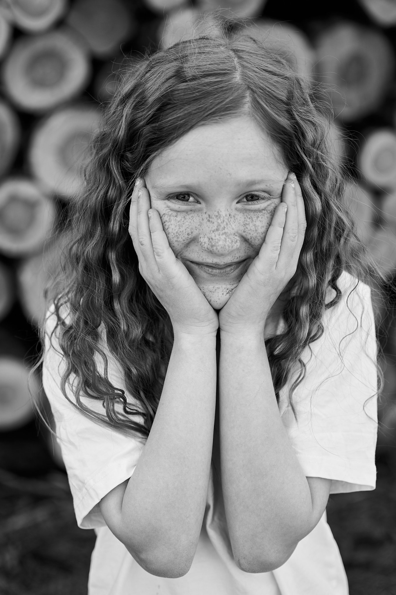En ung pige med krøllet hår og fregner, der smiler, mens hun holder ansigtet med begge hænder, foran en bunke af runde træstammer.