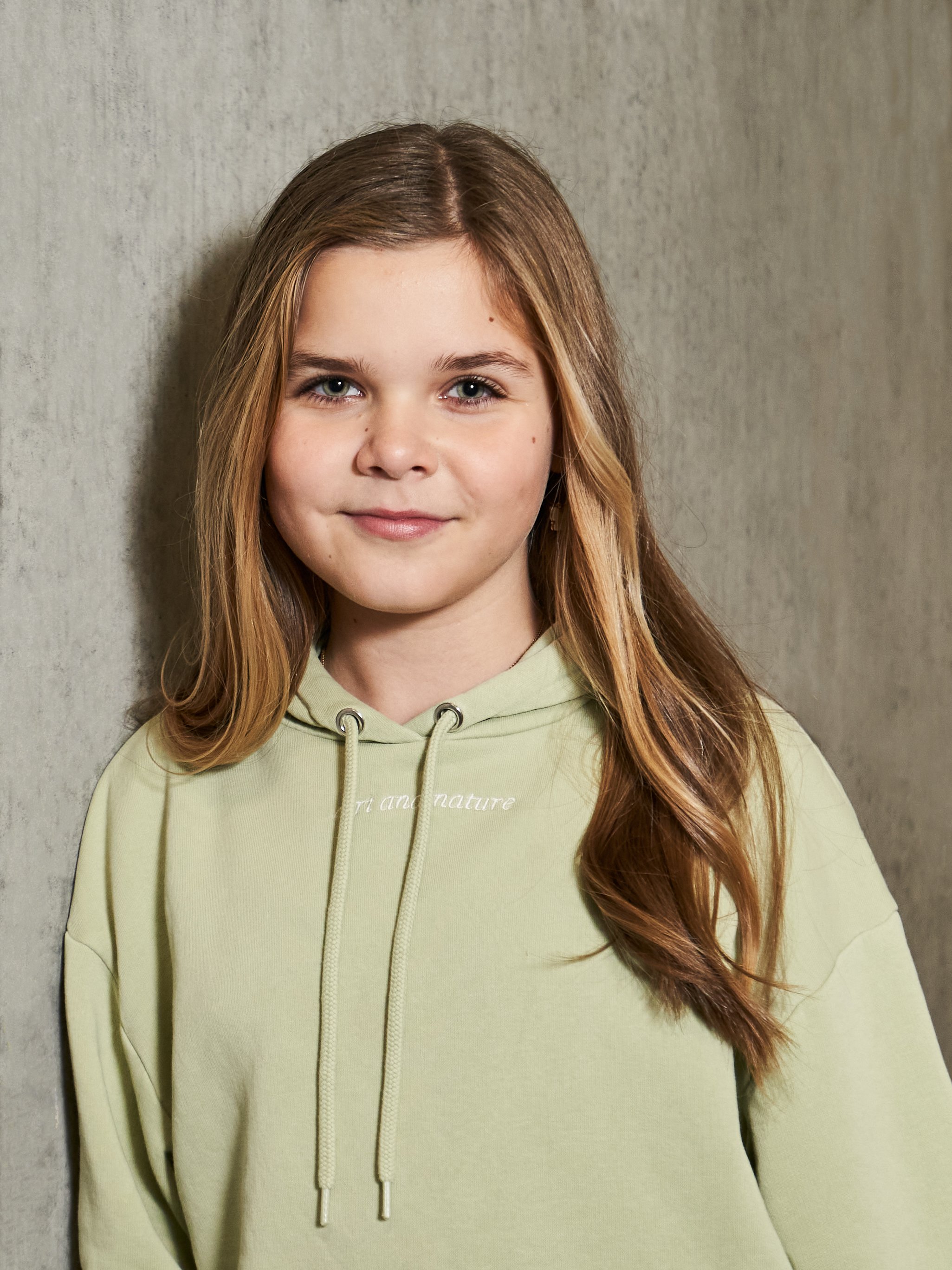 Portræt af en ung pige med brunt langt hår, iført lys gul hættetrøje, der står op ad en væg.