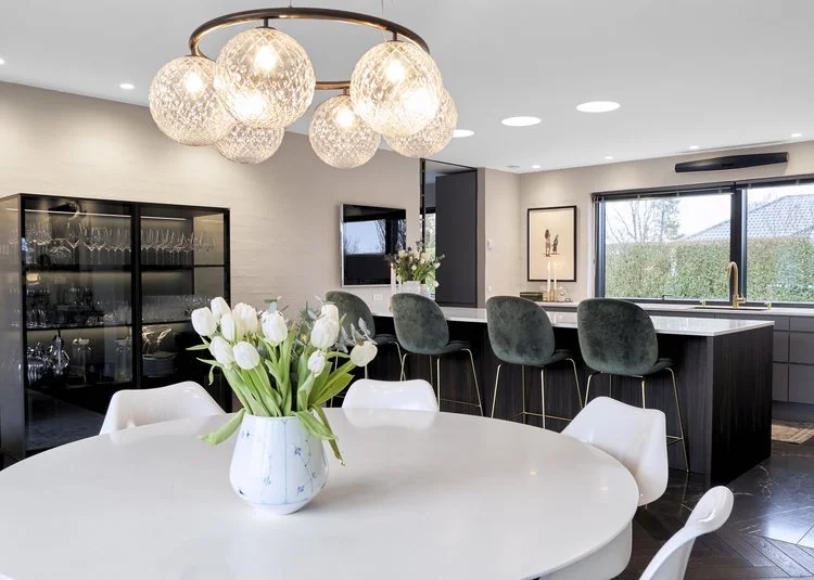 Moderne køkken- og diningområde med hvidt ovalbord med tulipaner, moderne stolre, bar med tre barstole, store vinduer og hungrende lyslamper.