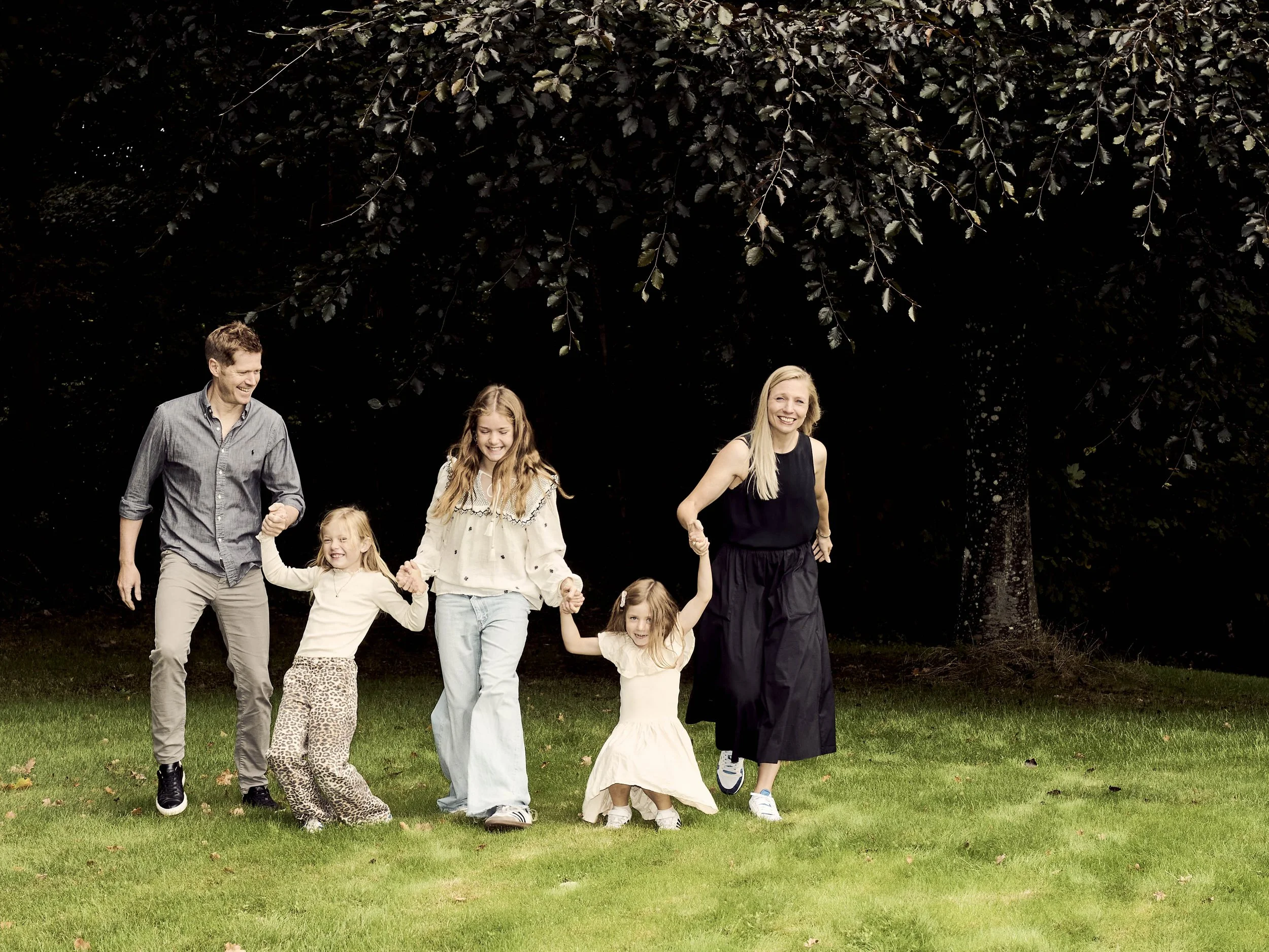 En gruppe mennesker går hånd i hånd på græs, foran store træer, smilende og glade