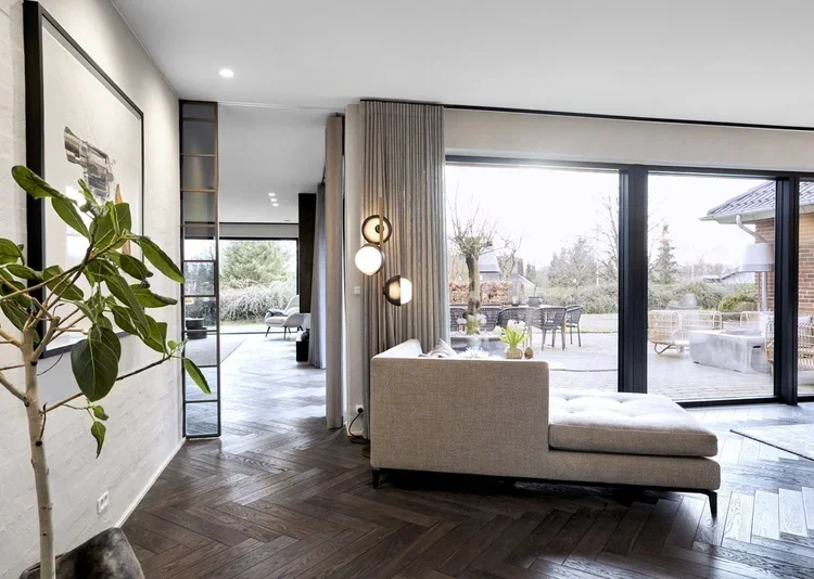 Moderne stue med stor vinduesfront, sofa, grøn plante, gulvlamper og udendørs terrasse med haveudstyr.