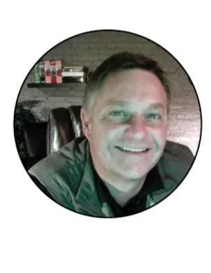 Smiling man sitting in a chair, in an indoor setting with shelves and boxes in the background.