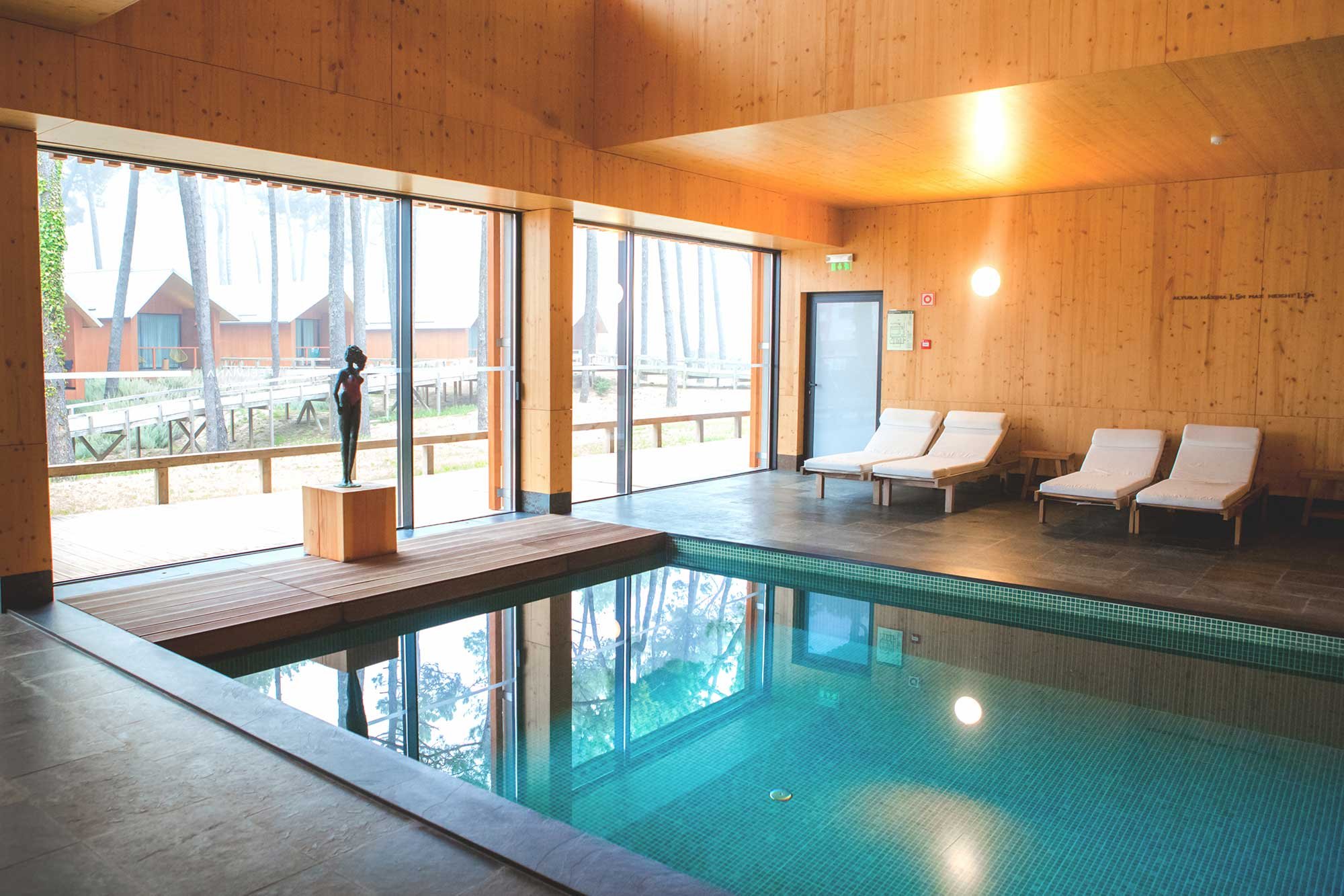 Indoor pool area with wooden walls and ceiling, large glass doors and windows looking outside at trees and cabins, three white lounge chairs, and a small statue on a wooden pedestal.