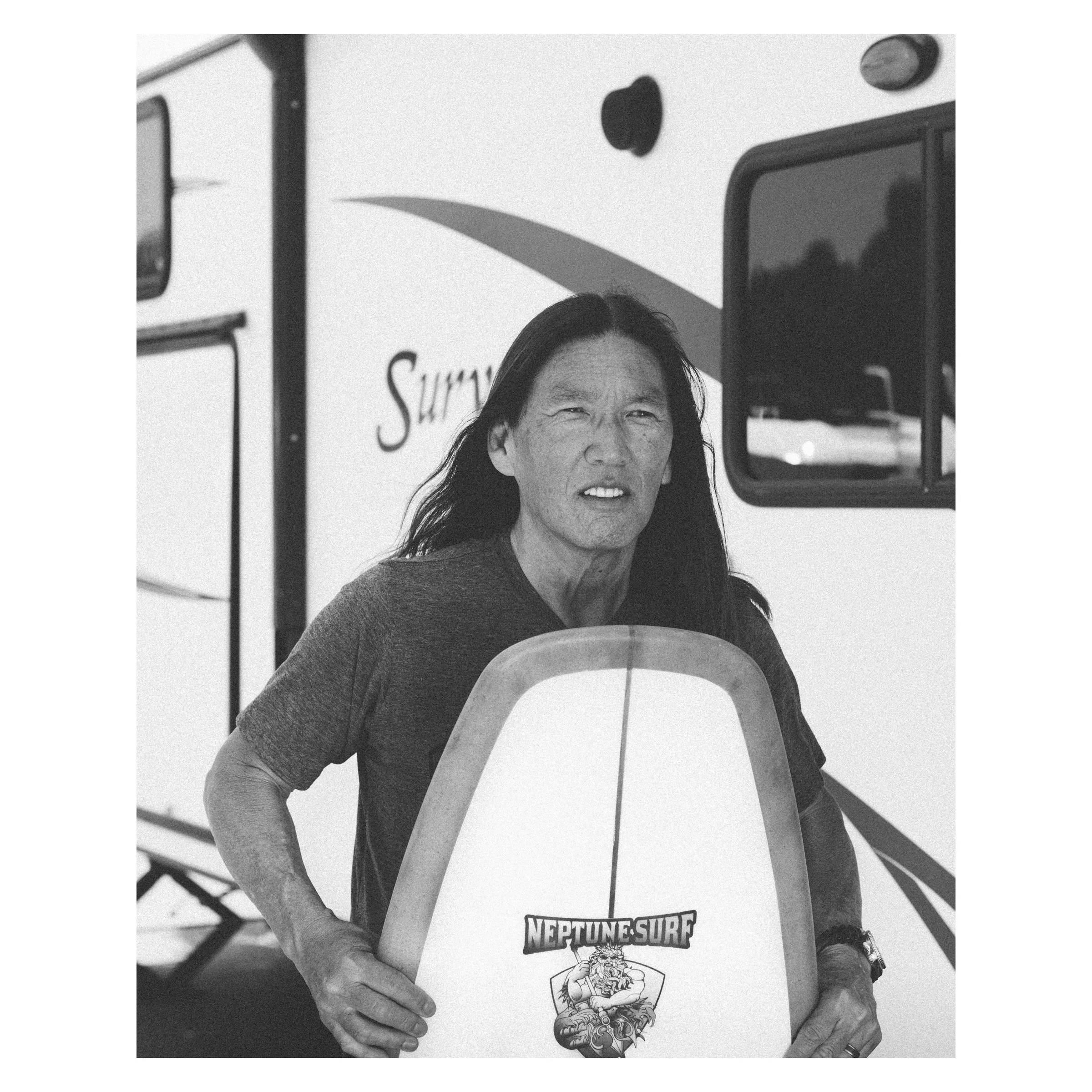 A person with long dark hair holding a surfboard in front of a camper trailer.