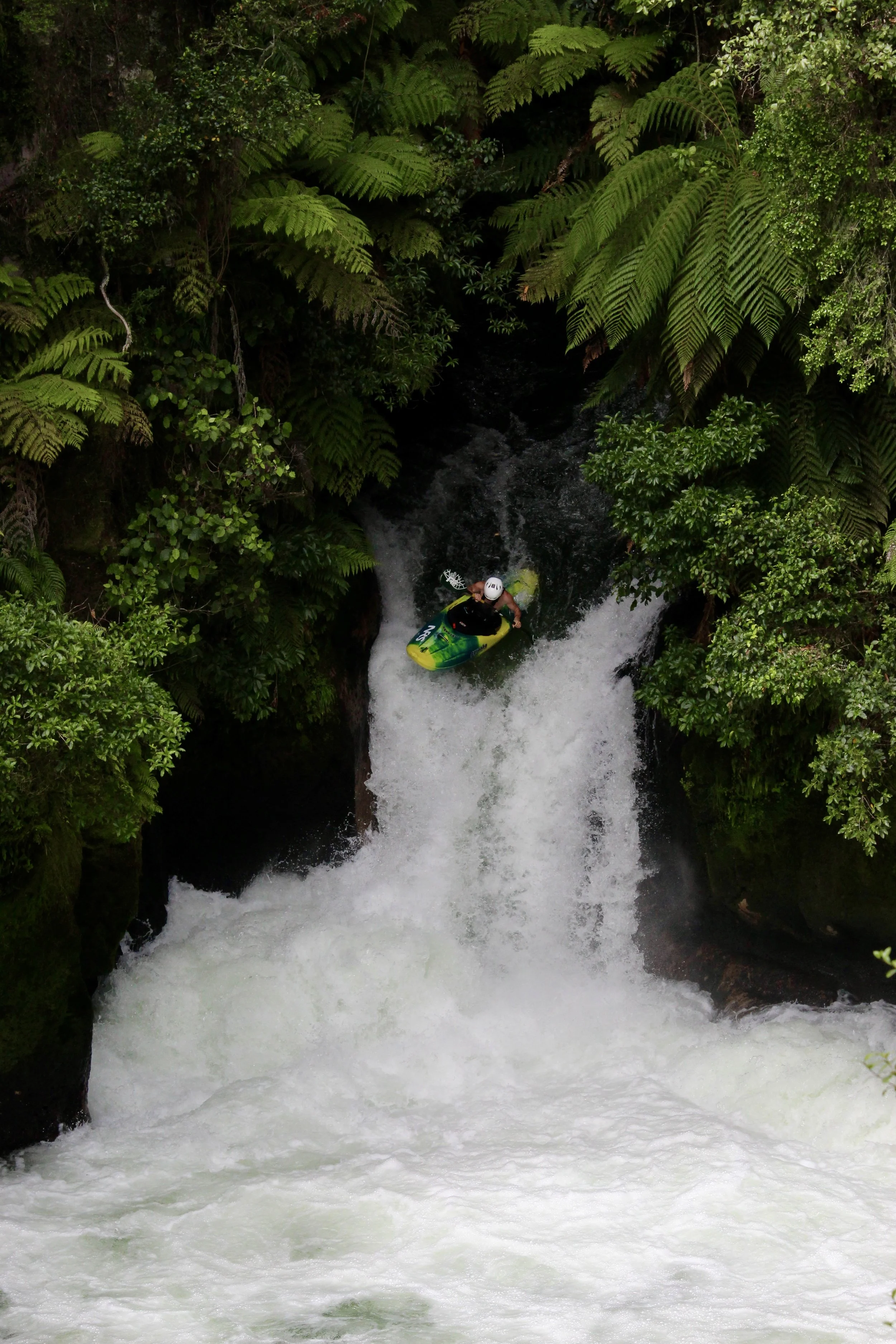 A man in outdoor gear holding a paddle, standing on a paved road with a hill and parked cars in the background.