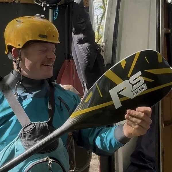 A man wearing a yellow helmet and blue jacket, sitting inside a vehicle, holding a paddle with a smiling expression.