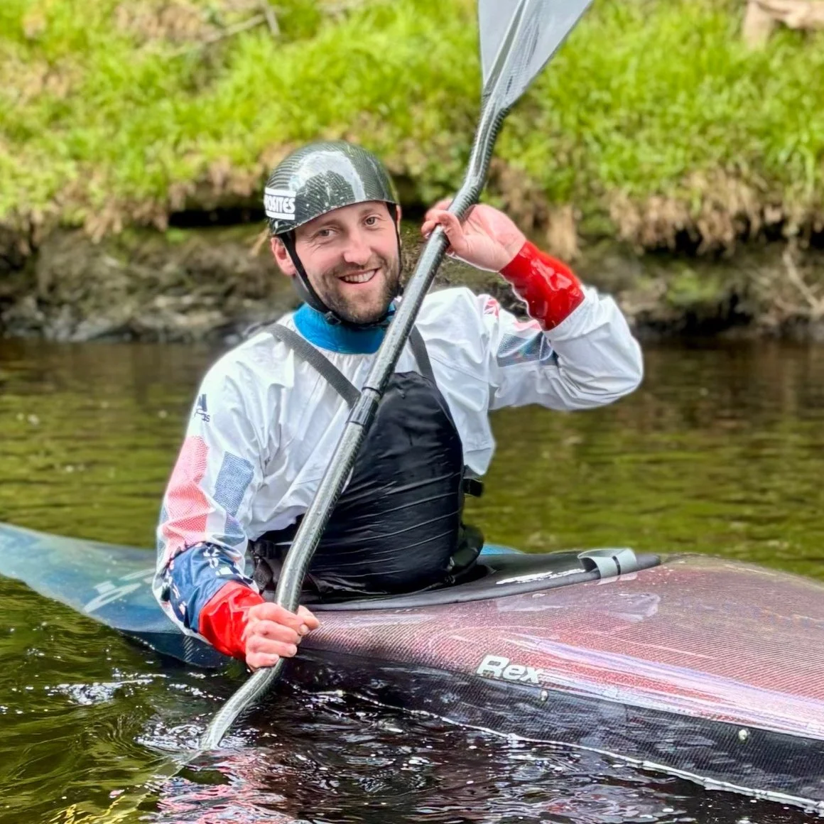 A man in a helmet and waterproof gear kayaking on a river, smiling and holding a paddle.