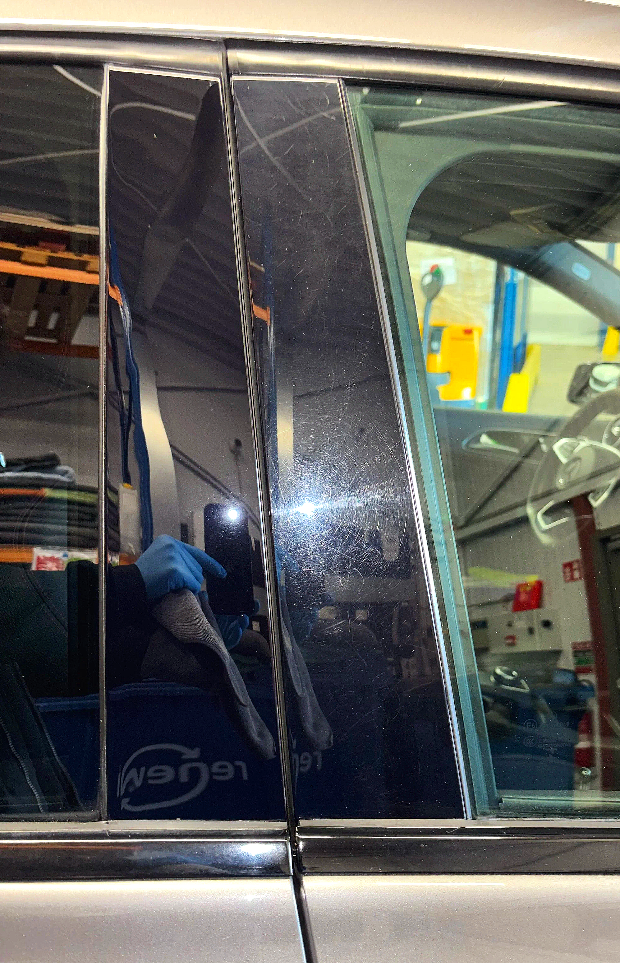 Close-up van een deel van een auto met reflecterende zwarte ramen en een kleine vertikale strip, zichtbaar in een werkplaats of garage.