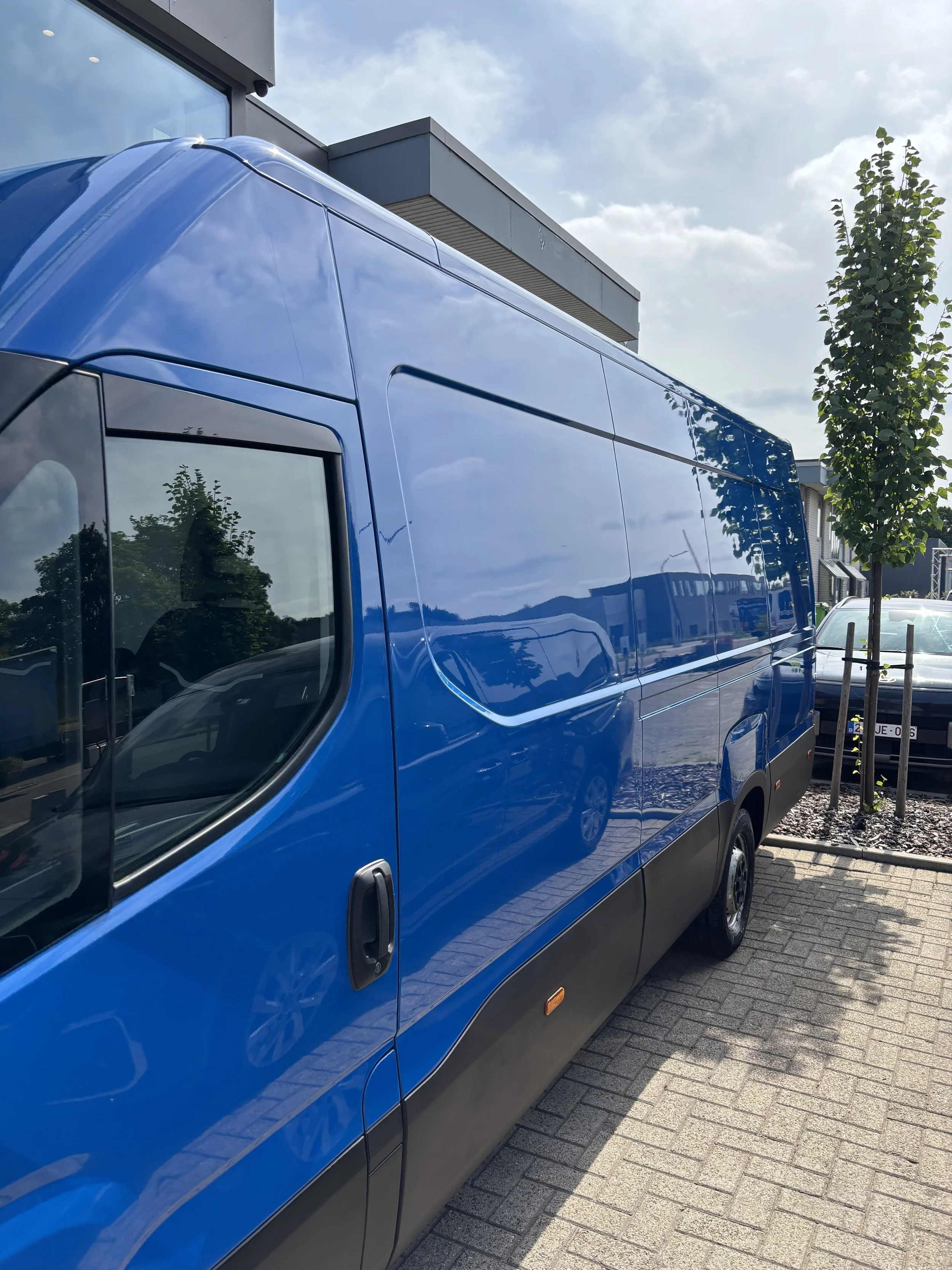 Blauwe bedrijfswagen geparkeerd op een oprit met een modern gebouw en bomen op de achtergrond onder een bewolkte hemel.