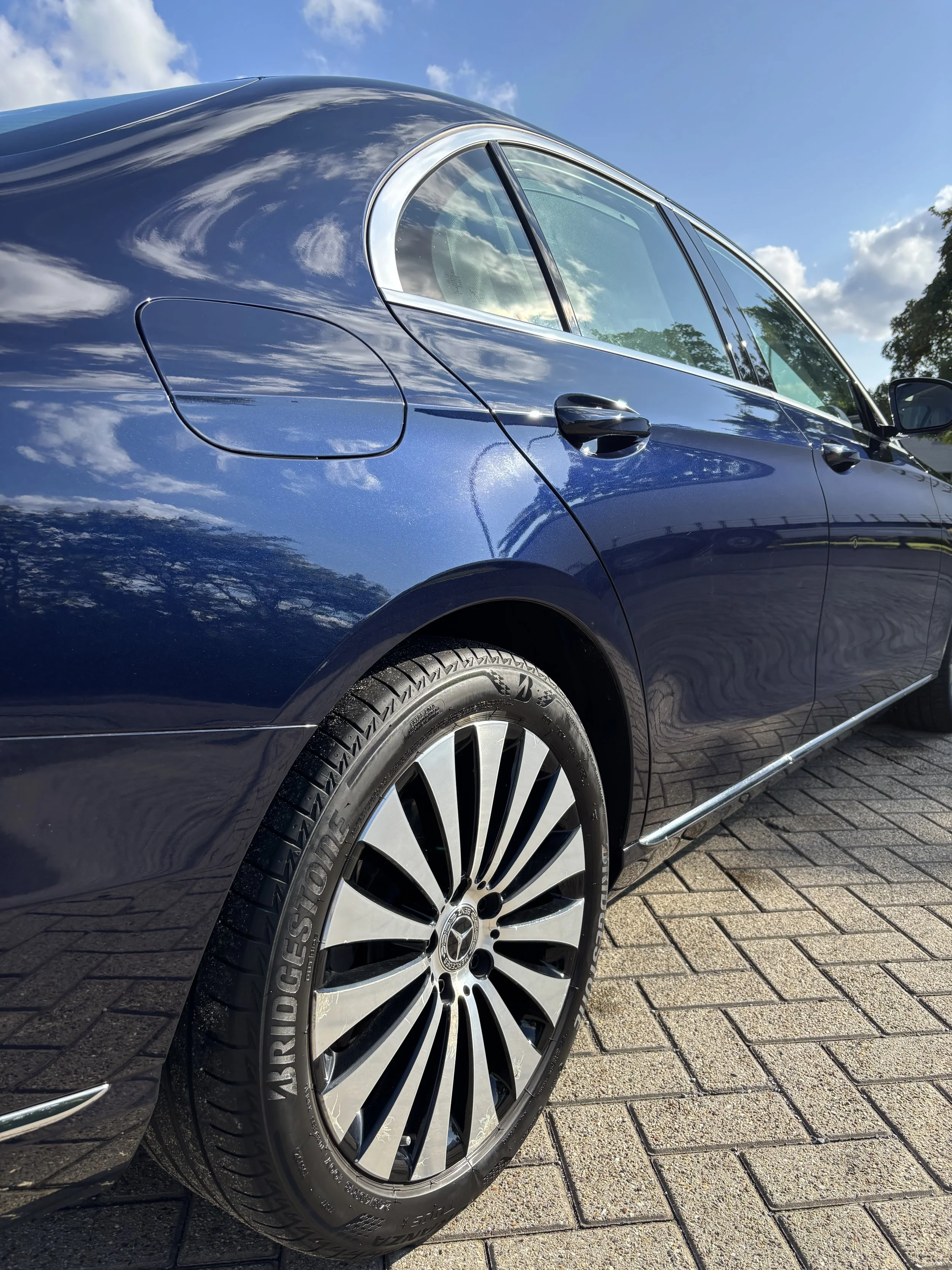 Zesdeurs blauwe Mercedes-Benz auto met plushen en spiegelreflecties, geparkeerd op stenen weg, onder een blauwe hemel met wolken en bomen op de achtergrond.
