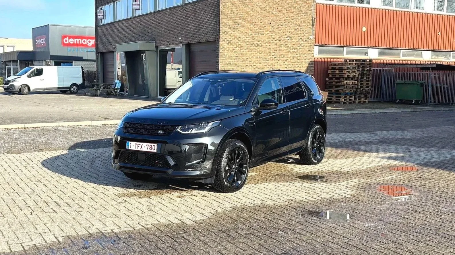 Zwarte SUV geparkeerd op een oprit met waterplassen eromheen, op de achtergrond gebouwen en een bestelwagen.