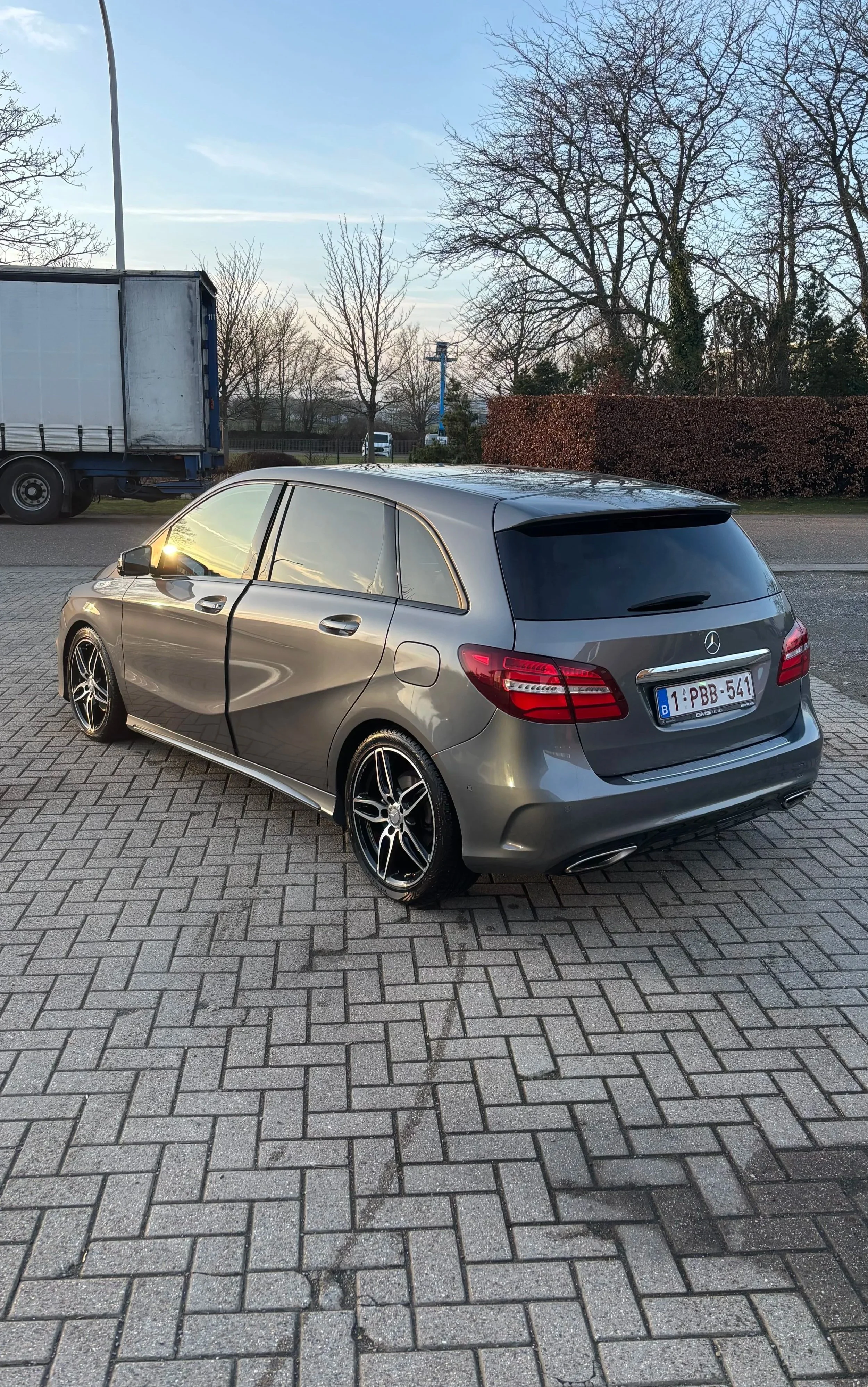 Een grijze Mercedes-Benz stationwagen geparkeerd op een betegeld parkeerterrein met bomen en een vrachtwagen op de achtergrond, onder een lichte hemel.