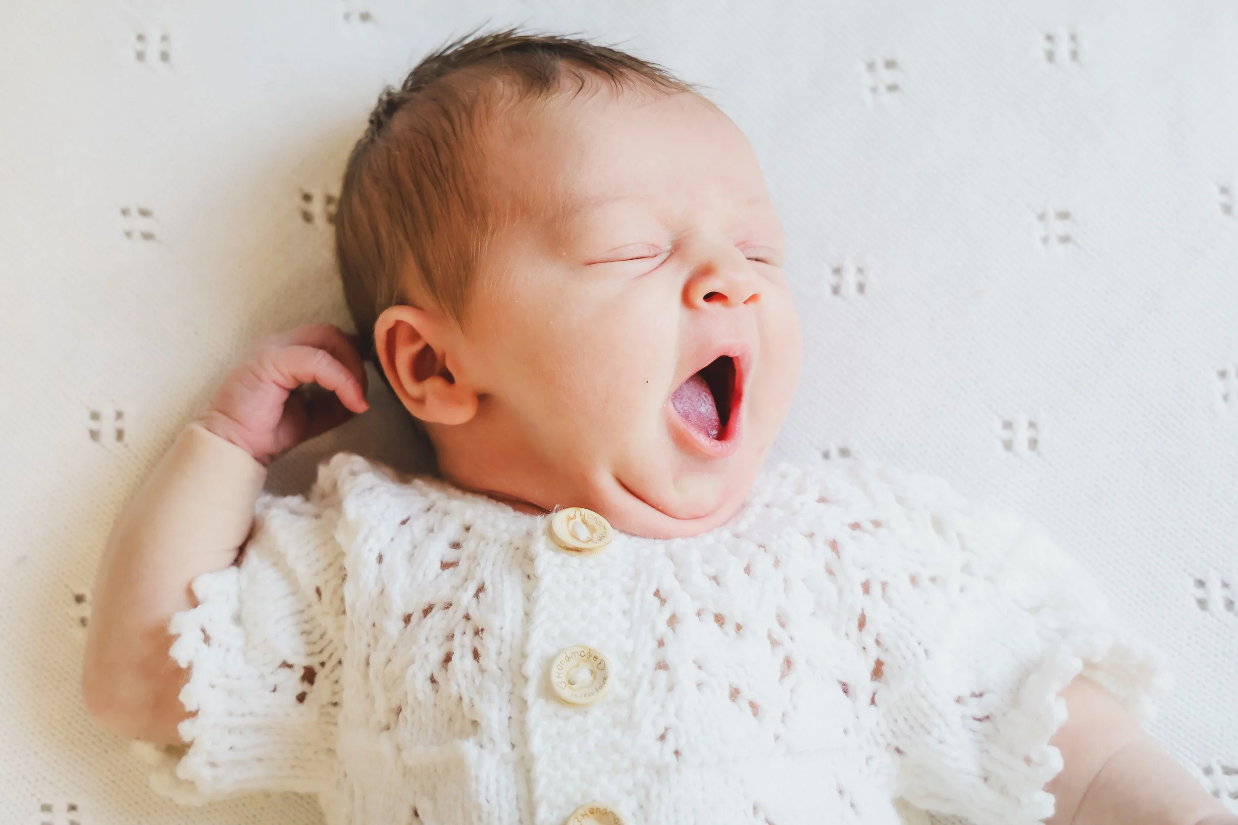 A newborn baby yawning while lying on a white textured blanket, wearing a white crocheted cardigan with wooden buttons.
