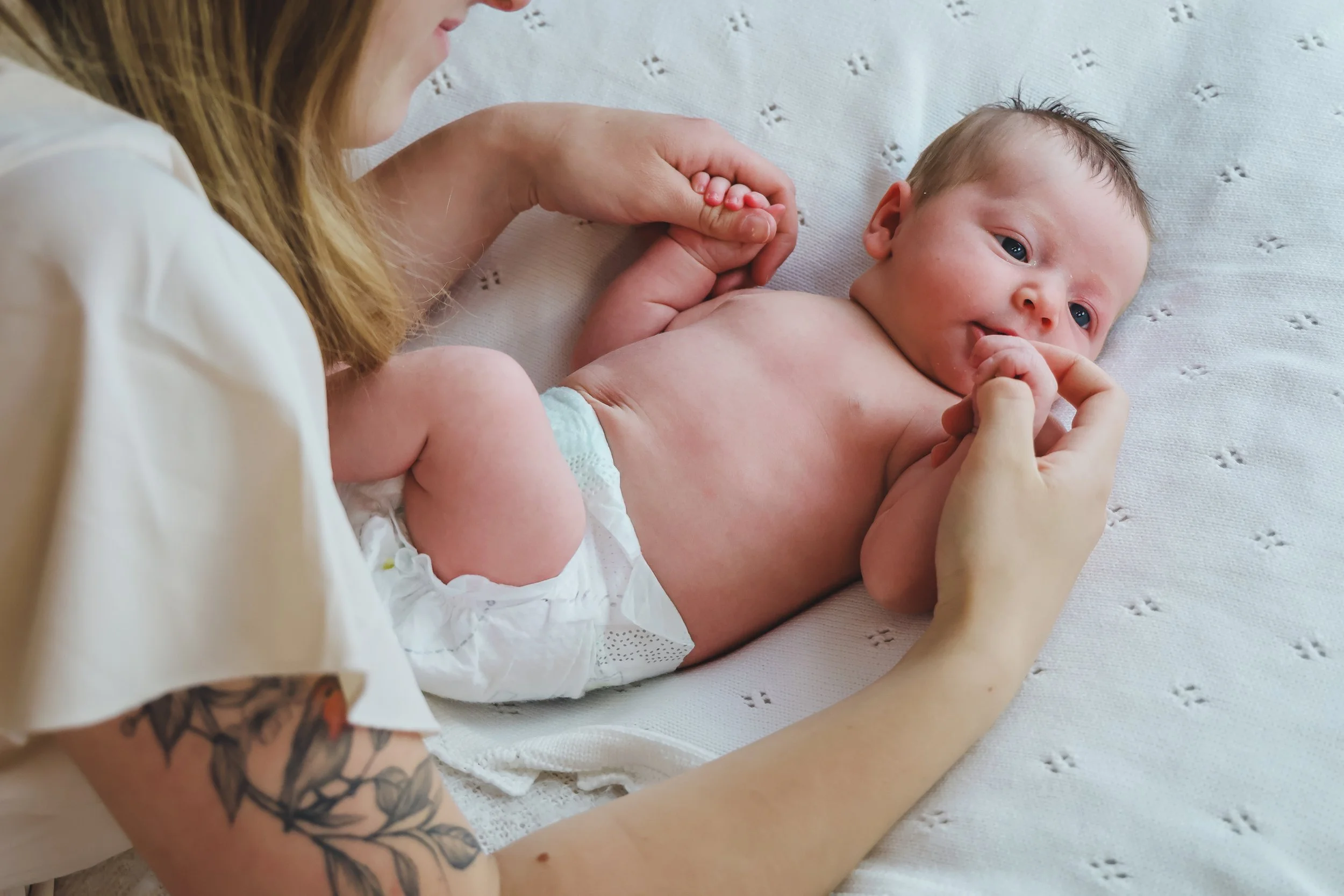 A woman with tattoos on her arm holding a baby, lying on a white textured blanket, looking at the camera and sucking her thumb.