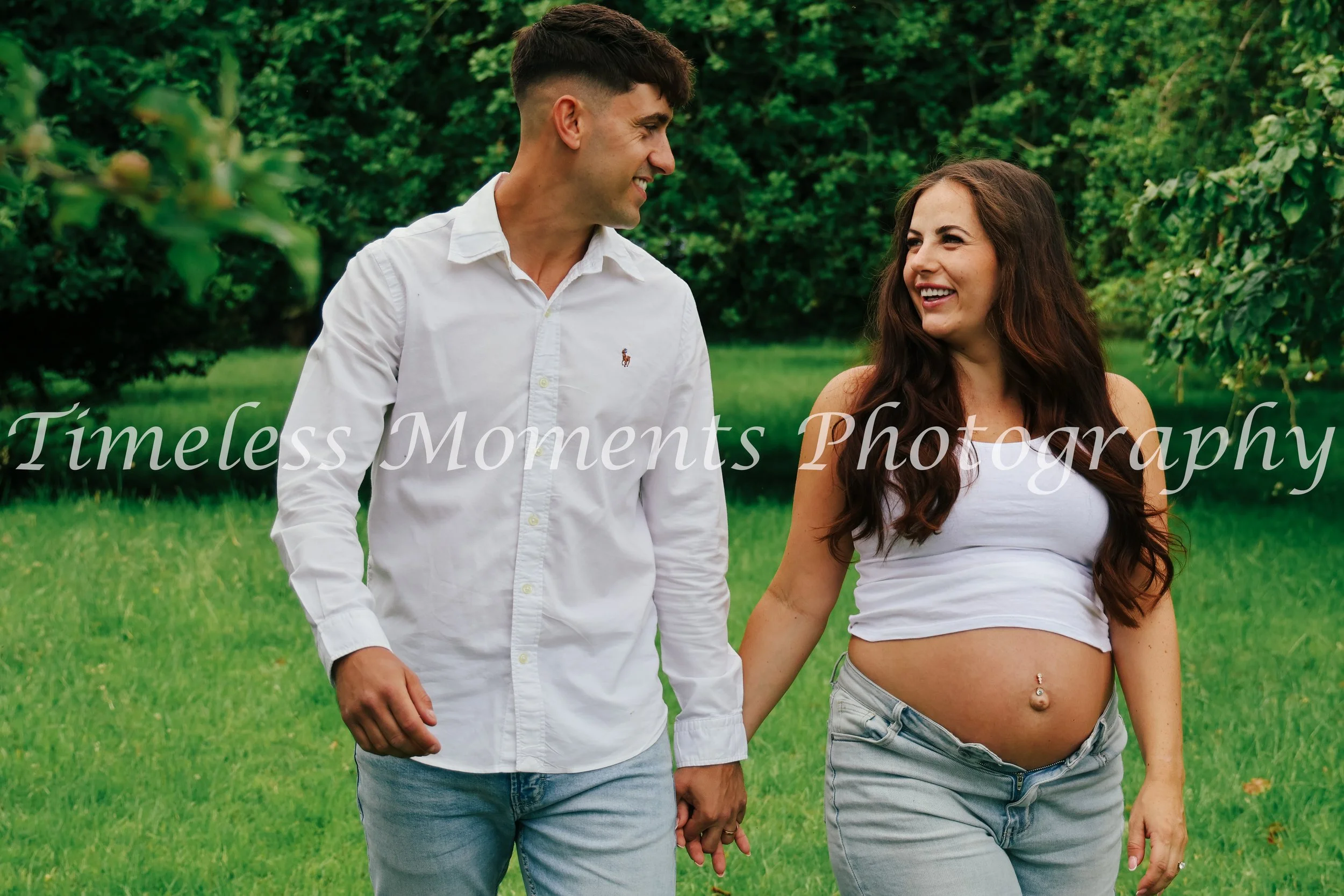 A smiling man and a pregnant woman walking hand in hand outdoors in a green park, enjoying a moment together.