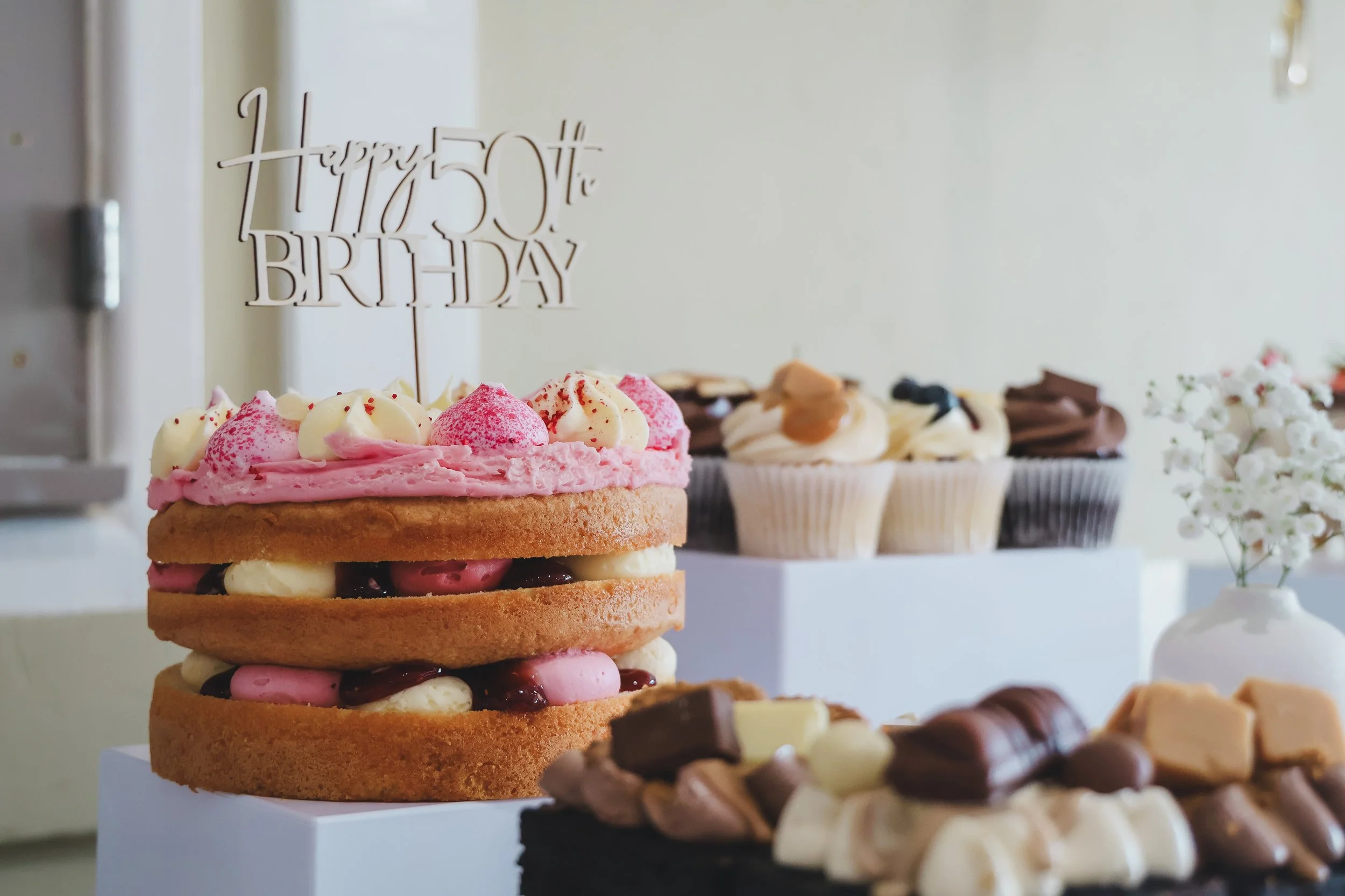 A layered cake with pink and white frosting and fruit toppings, celebrating a 50th birthday, with cupcakes and chocolates in the background.
