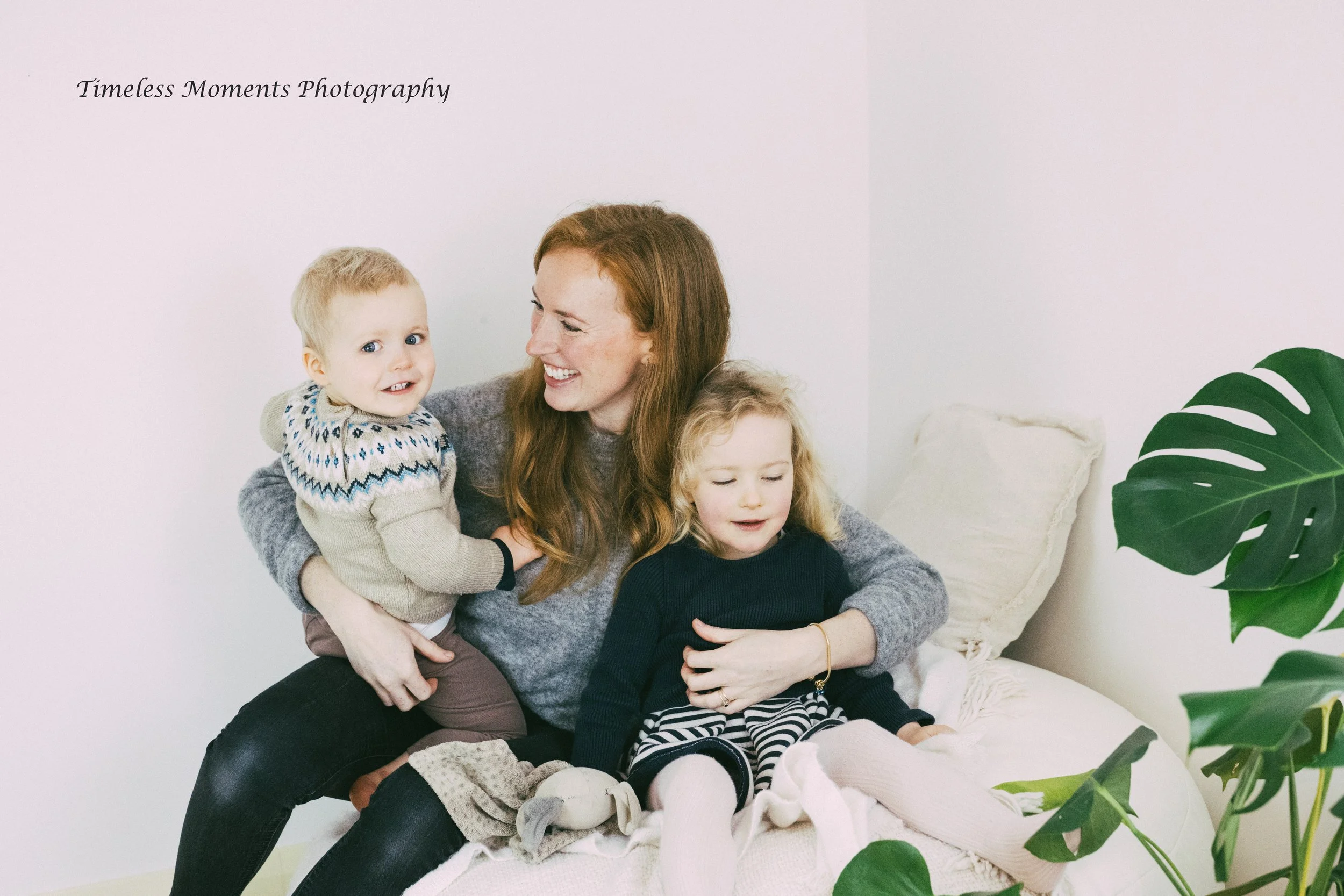 A woman with long red hair smiling while holding two young children, one on her lap and one standing beside her, sitting on a cream-colored chair in a bright room with a large green plant.