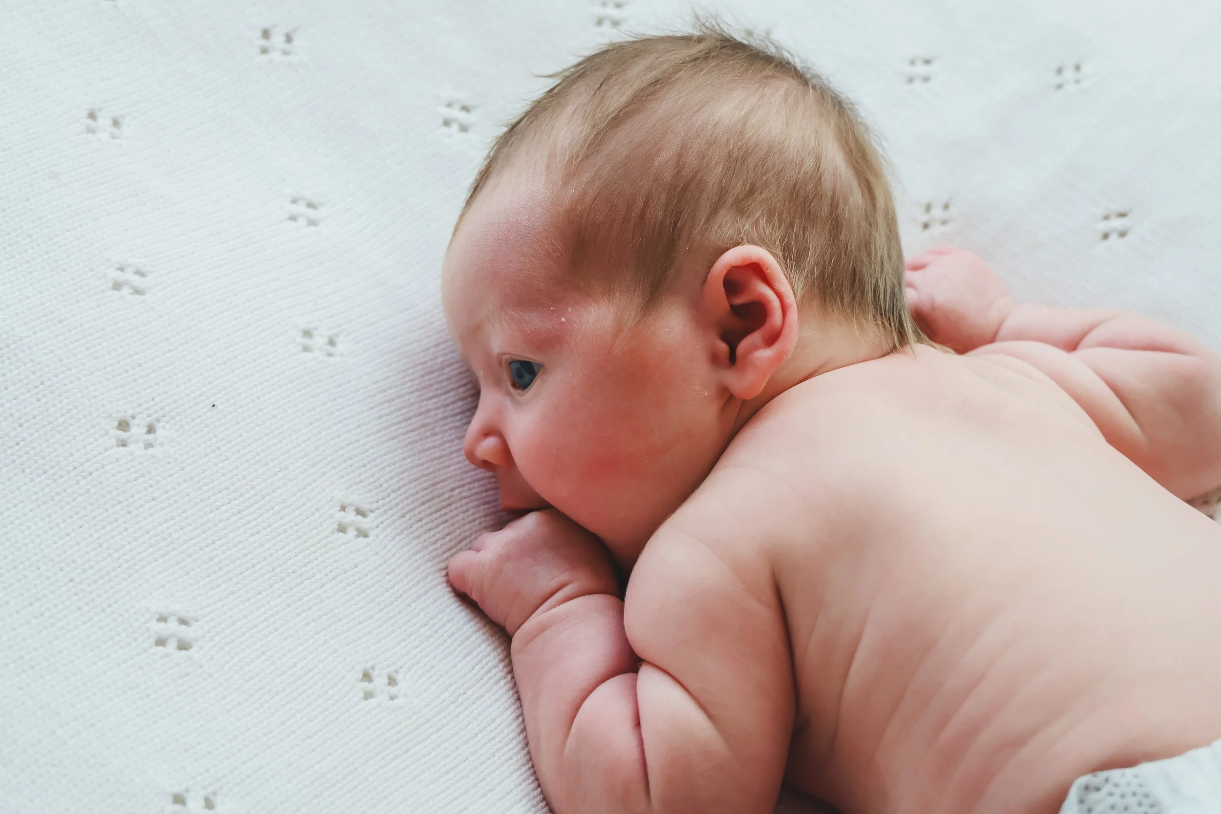 Close-up of a newborn baby lying on a white blanket with small cut-out star shapes, showing the baby's head, ear, and shoulder, with soft blonde hair and pinkish skin.
